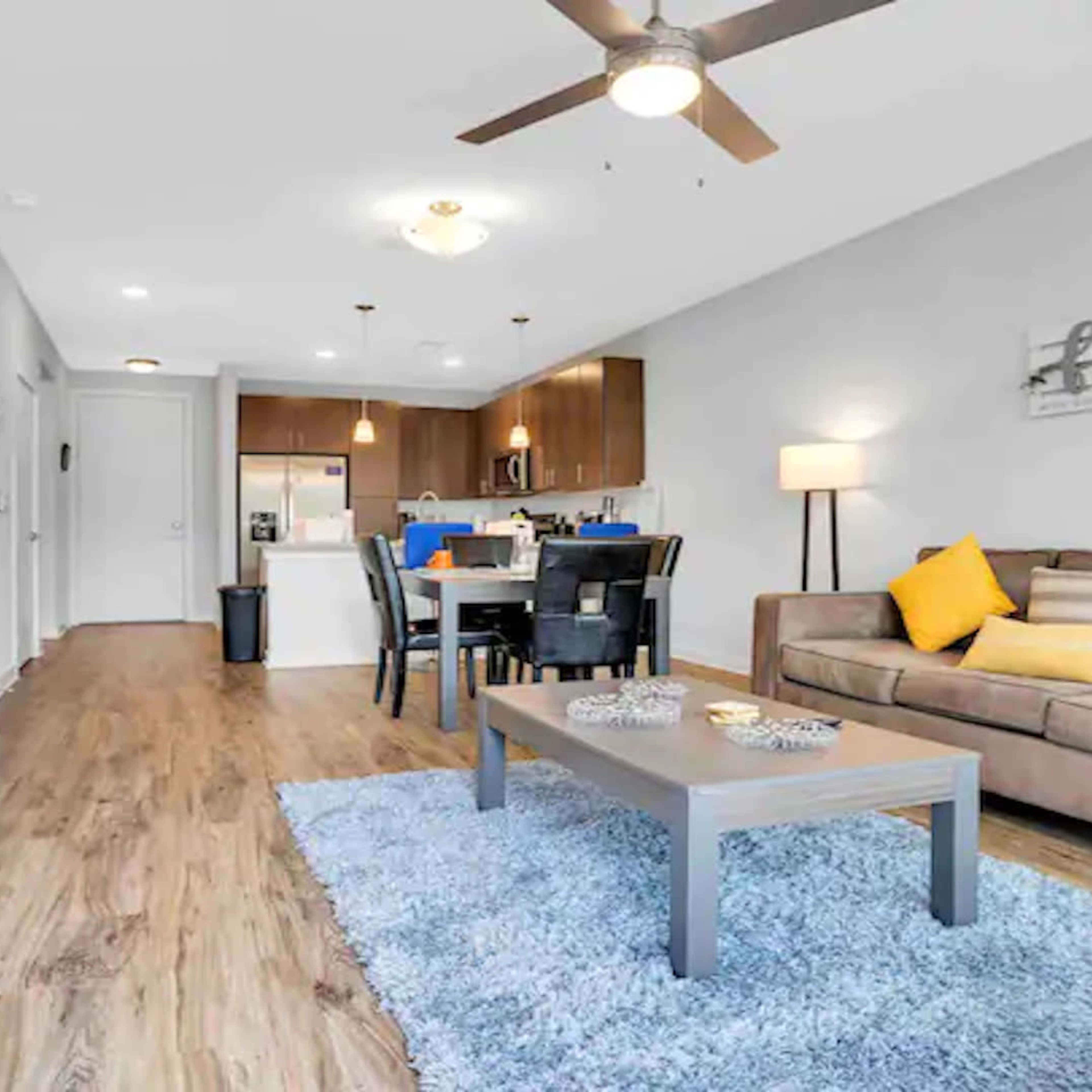 A modern living room with a brown sofa, a blue area rug, a dining table, and a kitchen in the background.