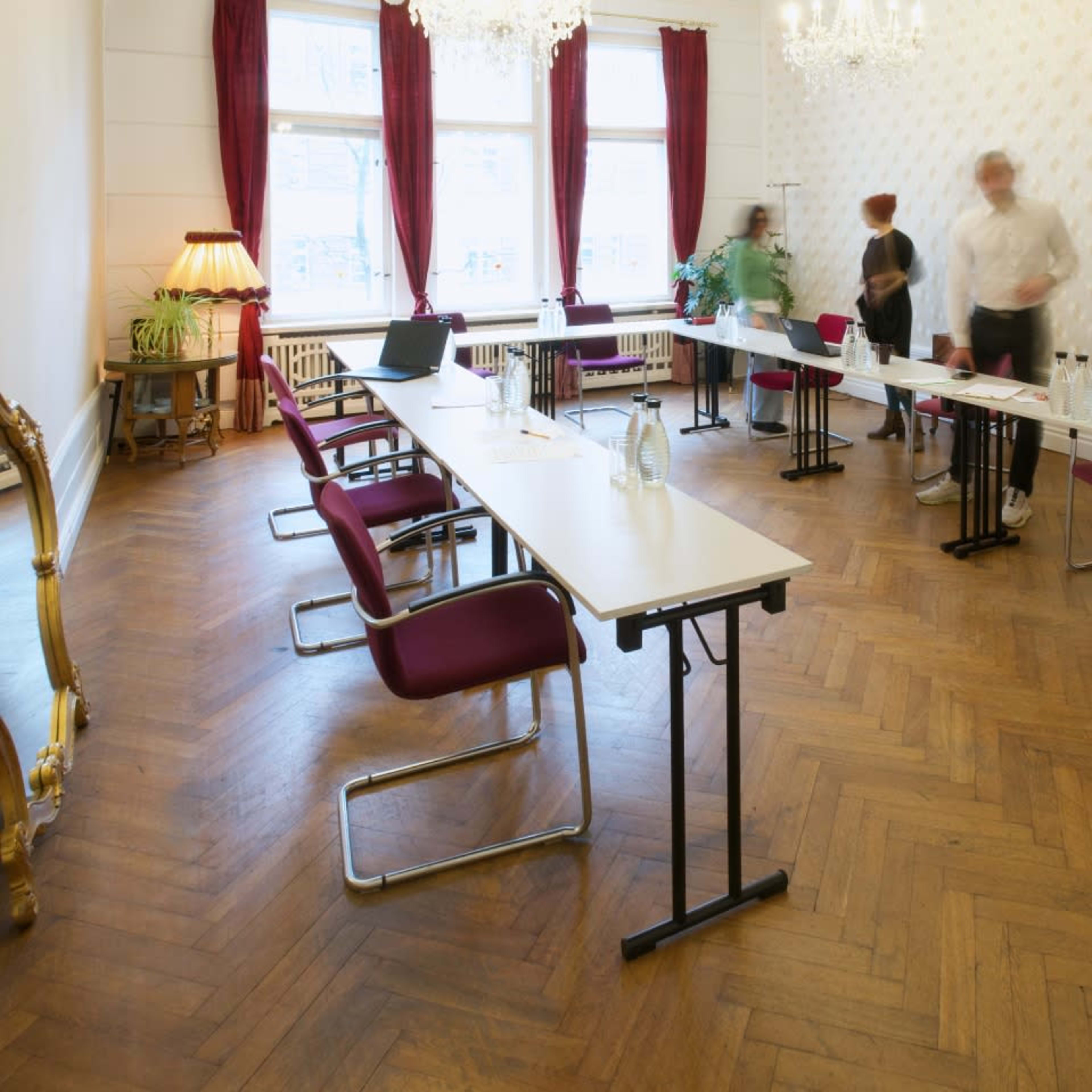 A meeting room with several tables and chairs arranged, featuring a window with red curtains and a chandelier overhead.
