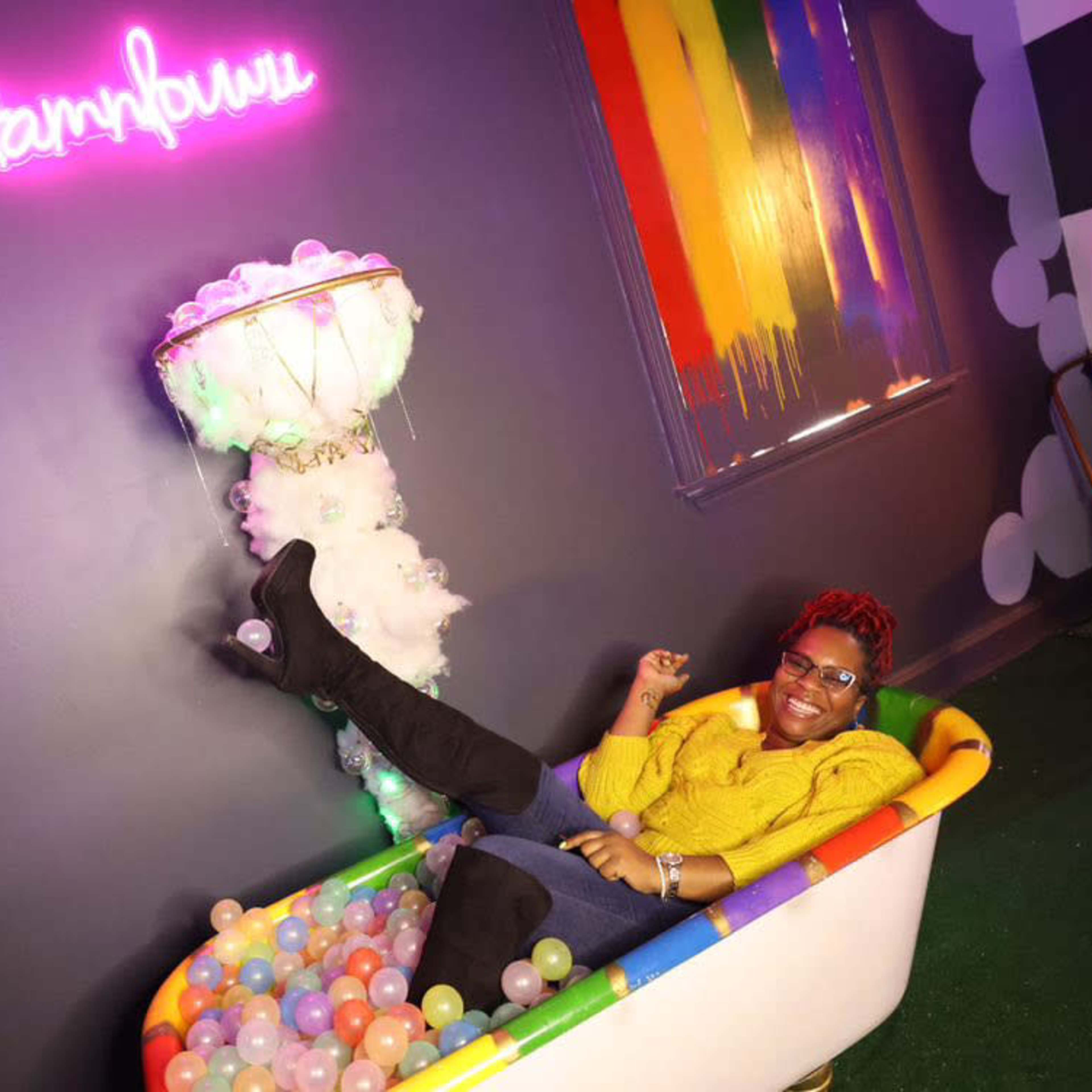 A person is sitting in a bathtub filled with colorful balls, with a neon sign above that reads "sampbulu" and a rainbow-striped wall in the background.
