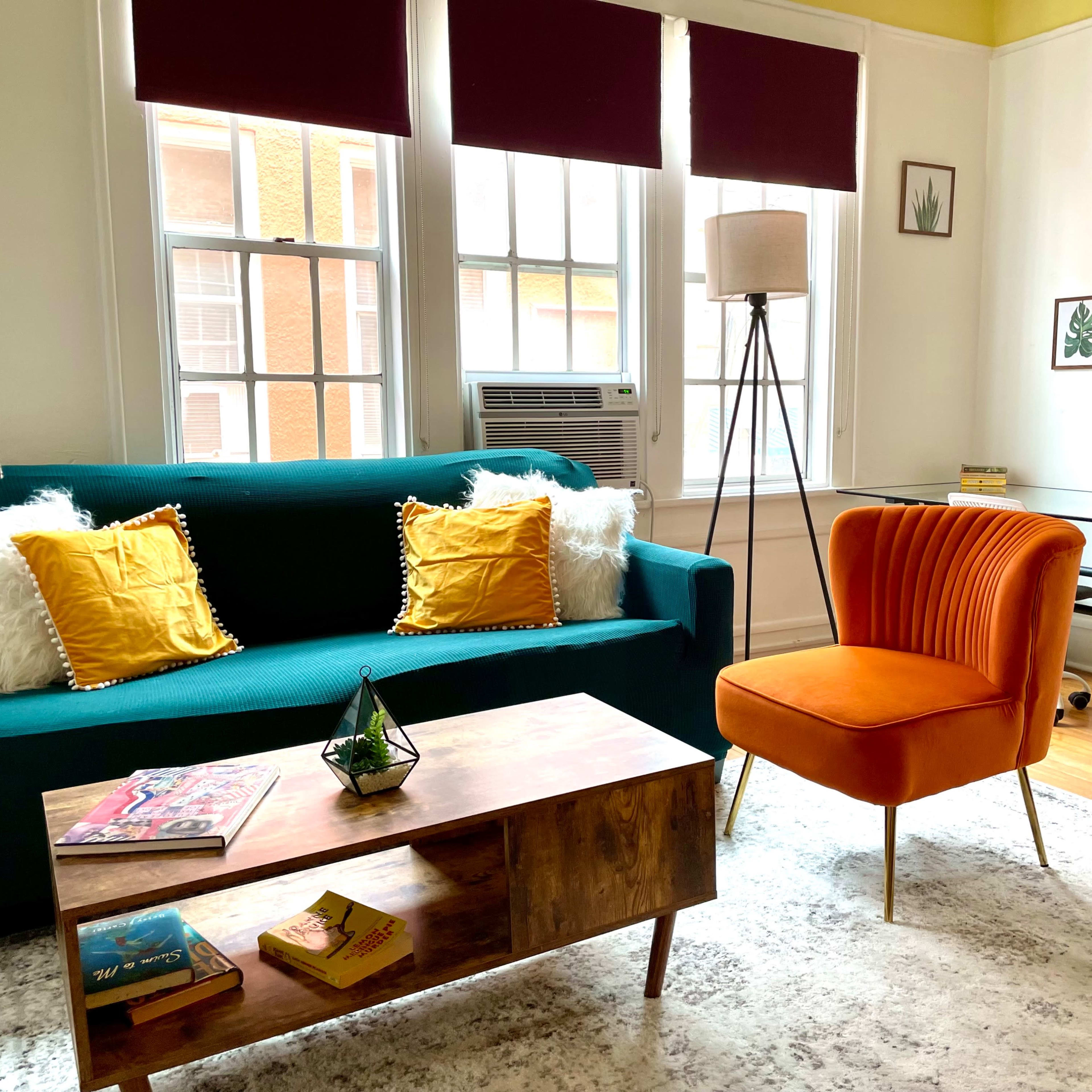 A cozy living room with a teal sofa, an orange accent chair, a wooden coffee table, and large windows letting in natural light.