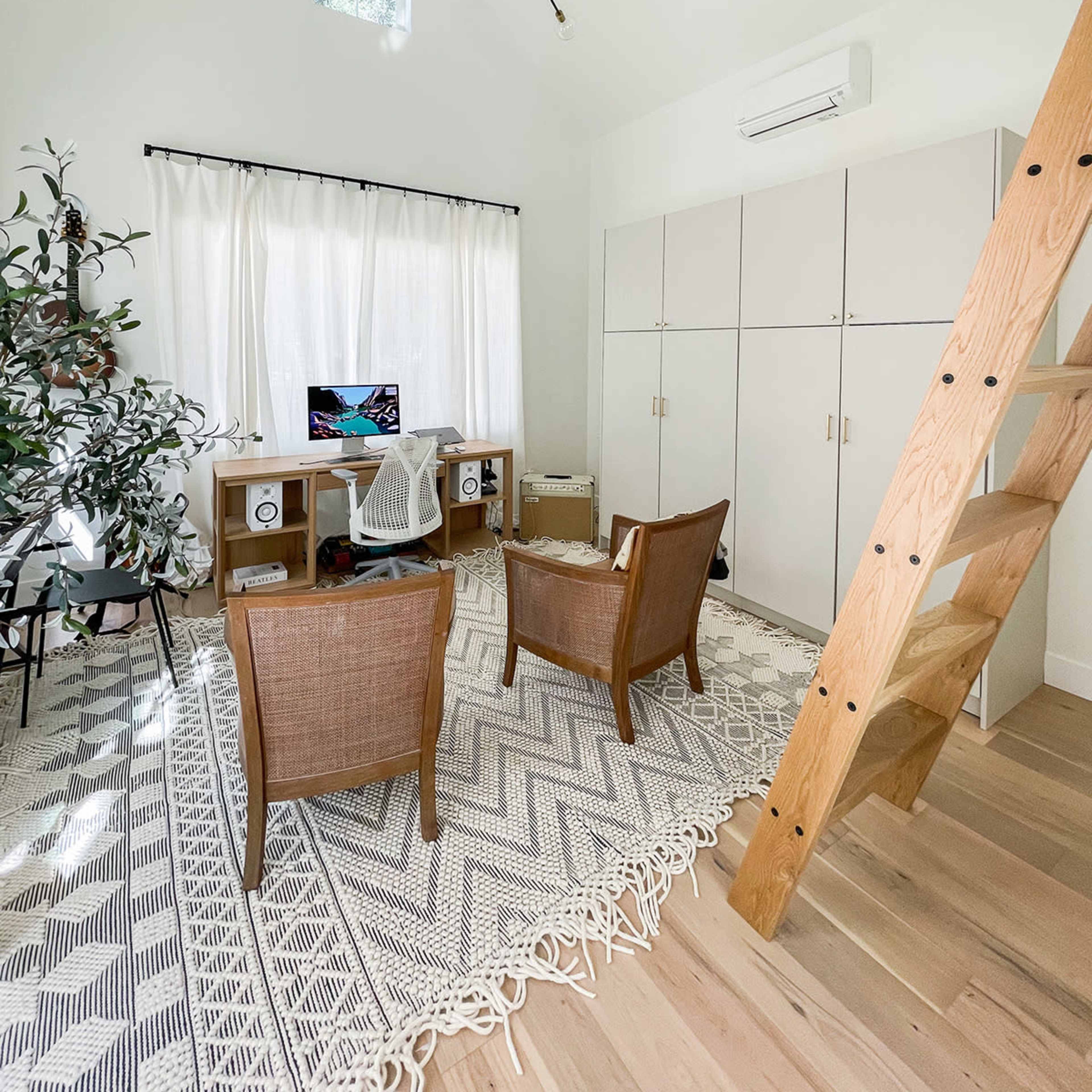 A bright, minimalist home office with two wicker chairs, a desk with a computer, and a staircase leading to a loft area.