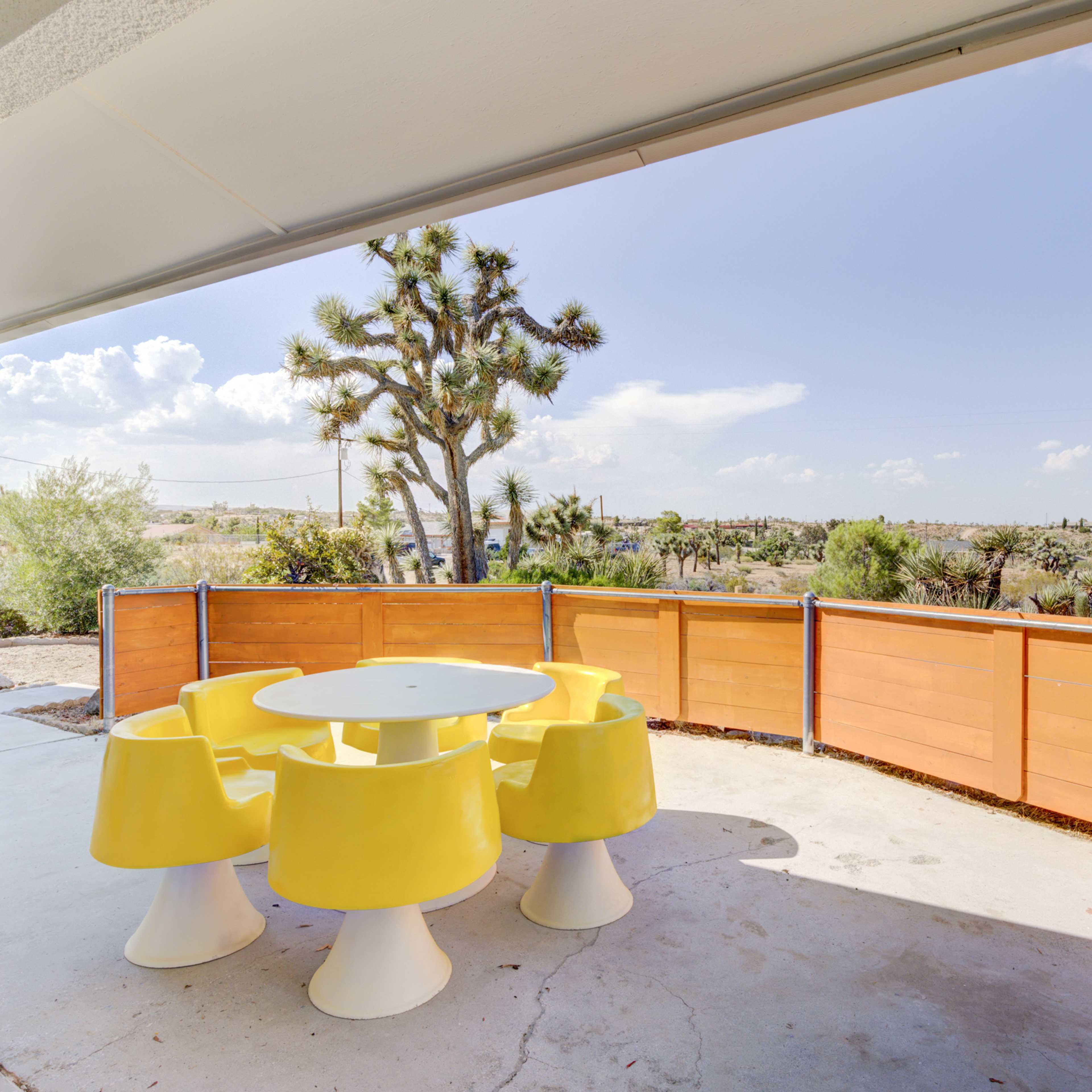 A patio area features a round white table surrounded by yellow chairs, with a desert landscape and scattered trees in the background.