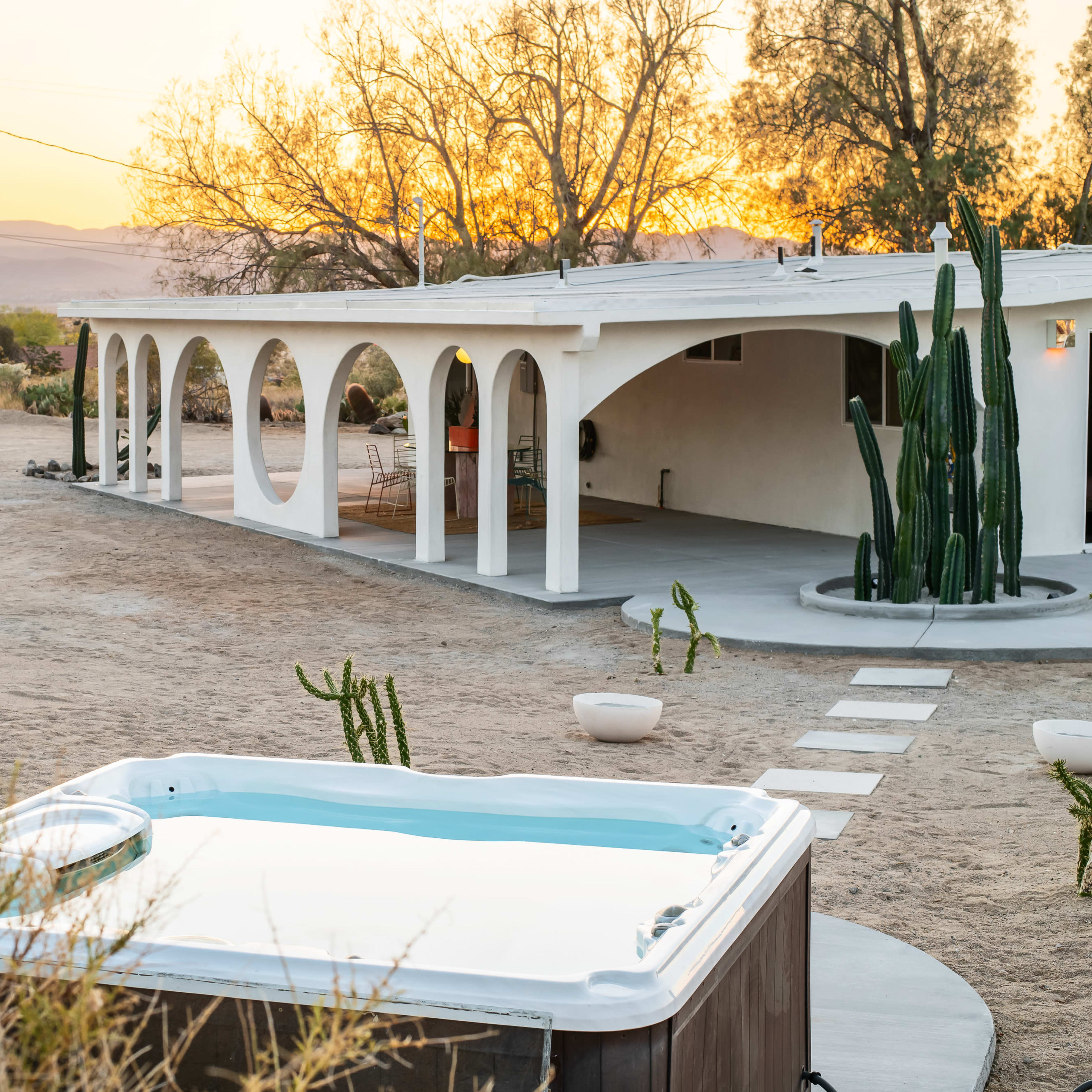 A modern home with an archway features a hot tub in the foreground, surrounded by desert landscaping at sunset.