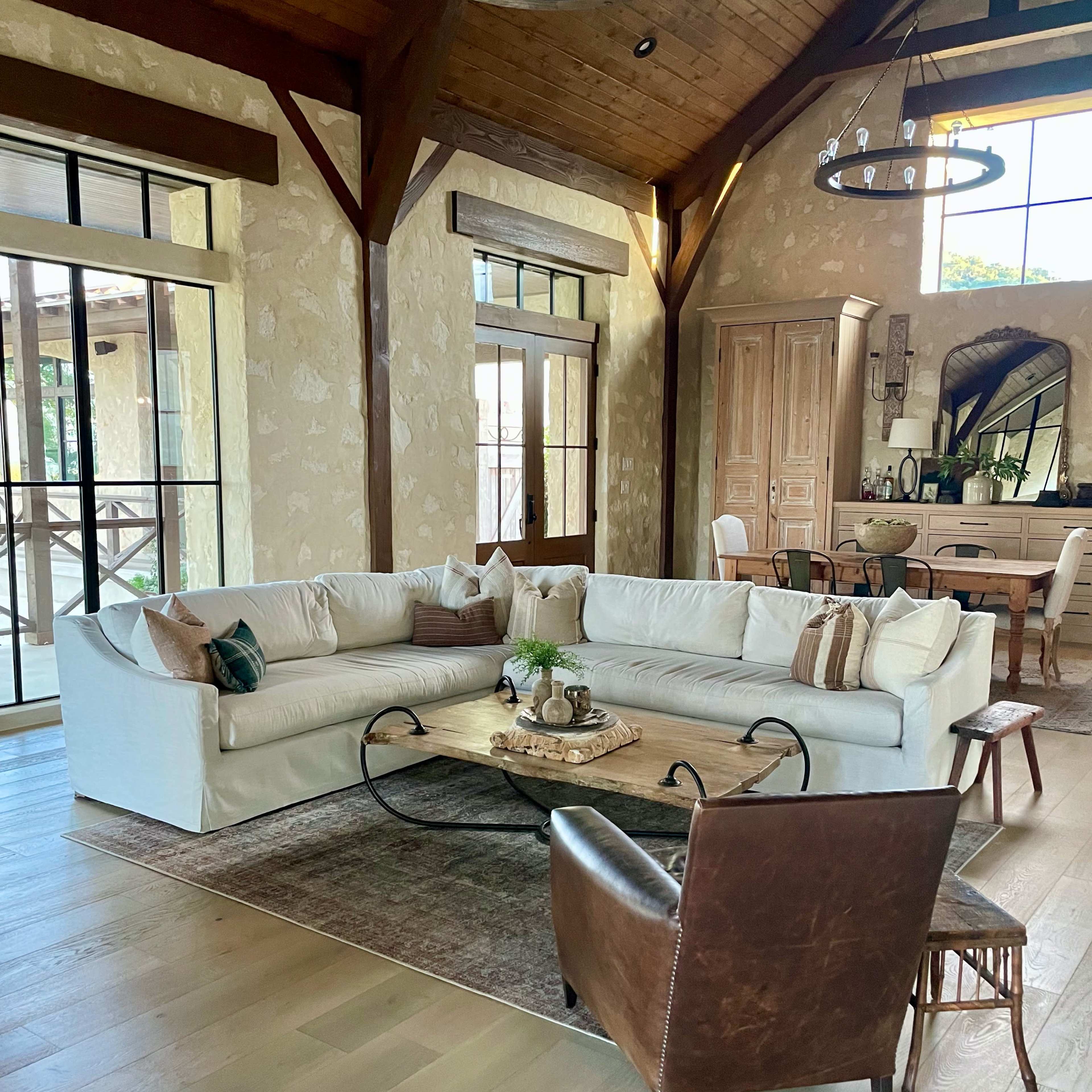 A spacious living room featuring a large white sectional sofa, a circular wooden coffee table, and a brown leather armchair, all set against a backdrop of wooden beams and large windows.