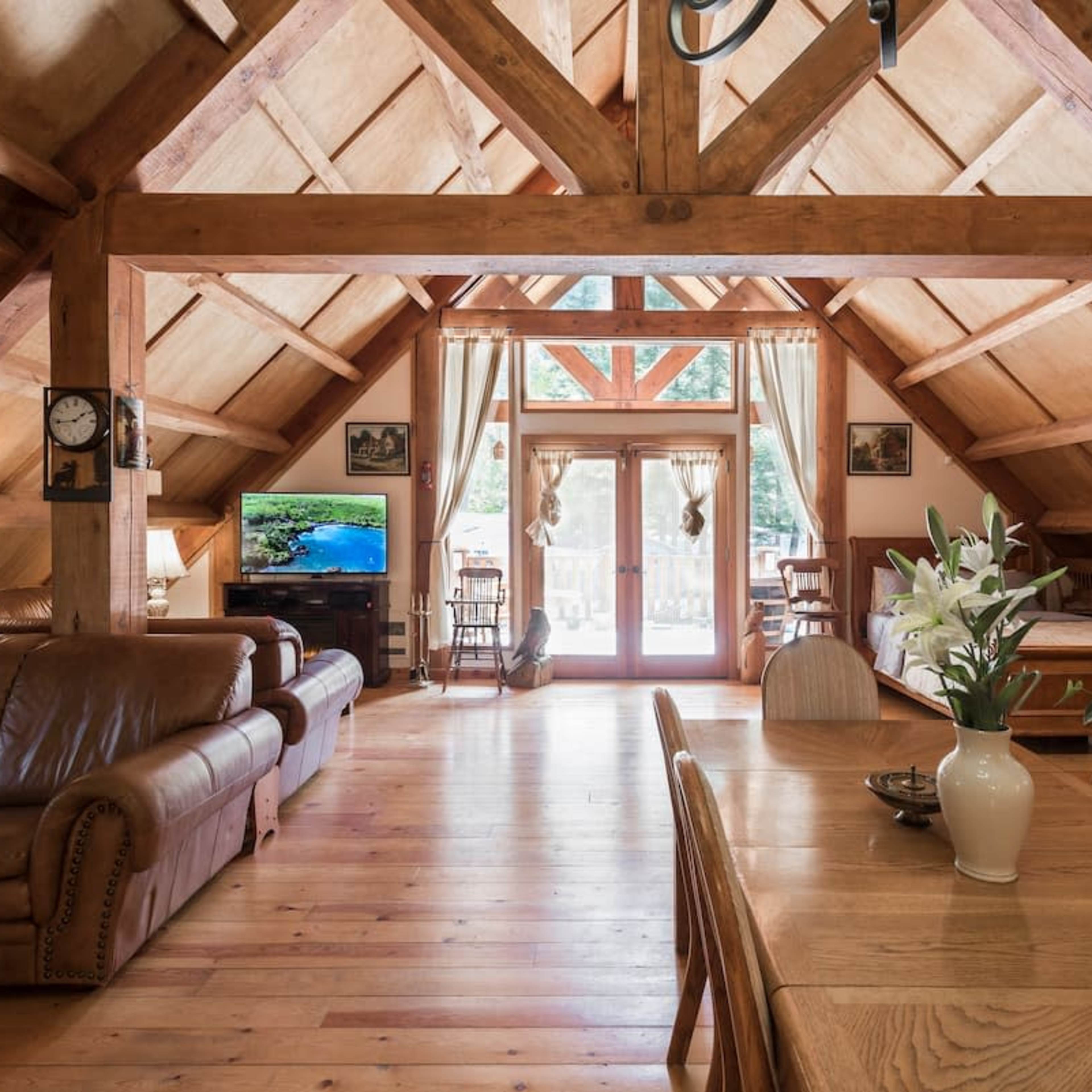 A spacious, wood-beamed interior of a cozy living area with a dining table, a leather couch, and large windows opening to a deck.