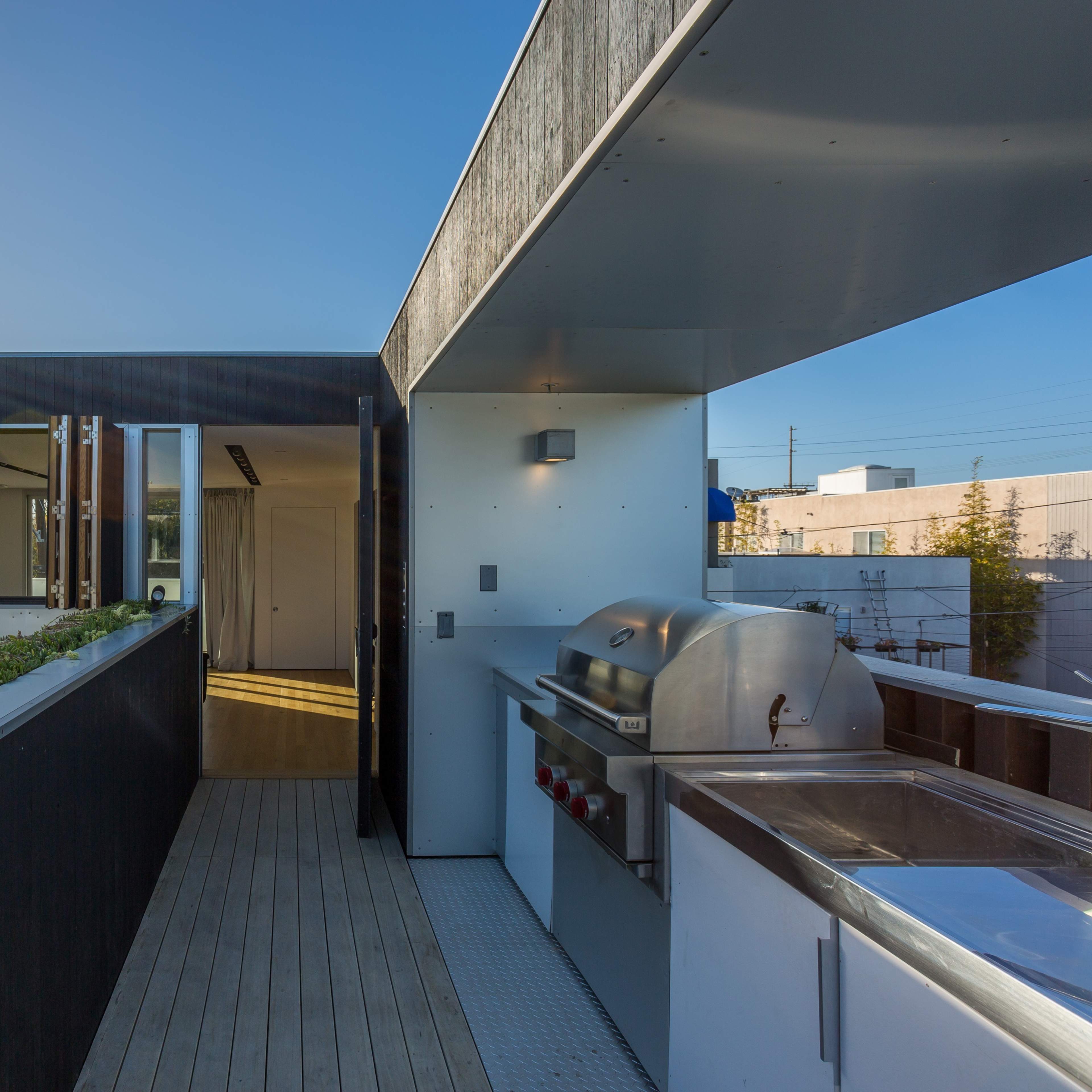 A modern rooftop terrace with a stainless steel grill, surrounded by planters, featuring glass walls and a view of neighboring buildings.