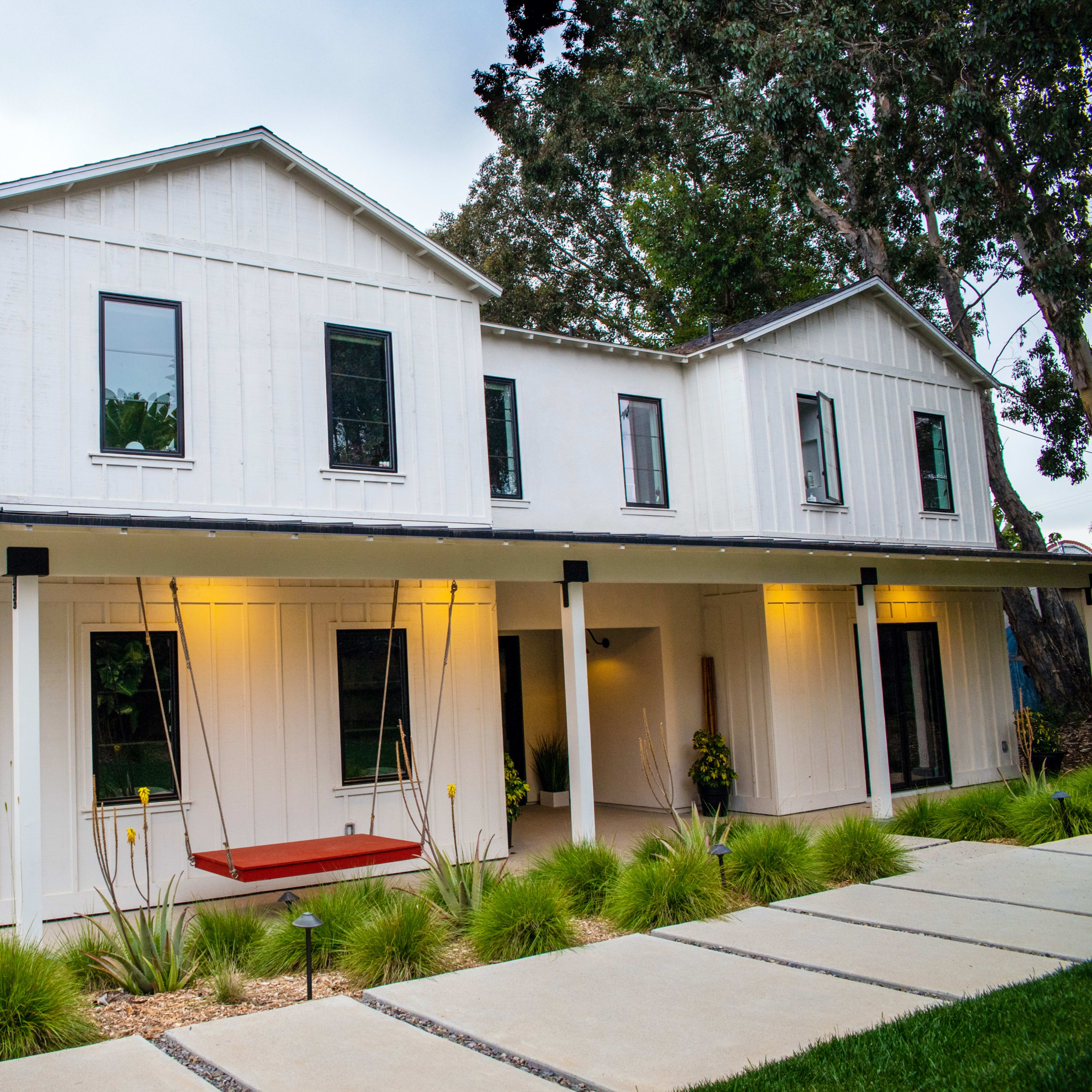 A modern, two-story white house with black windows and a porch featuring a red swing, surrounded by landscaped greenery and a pathway.