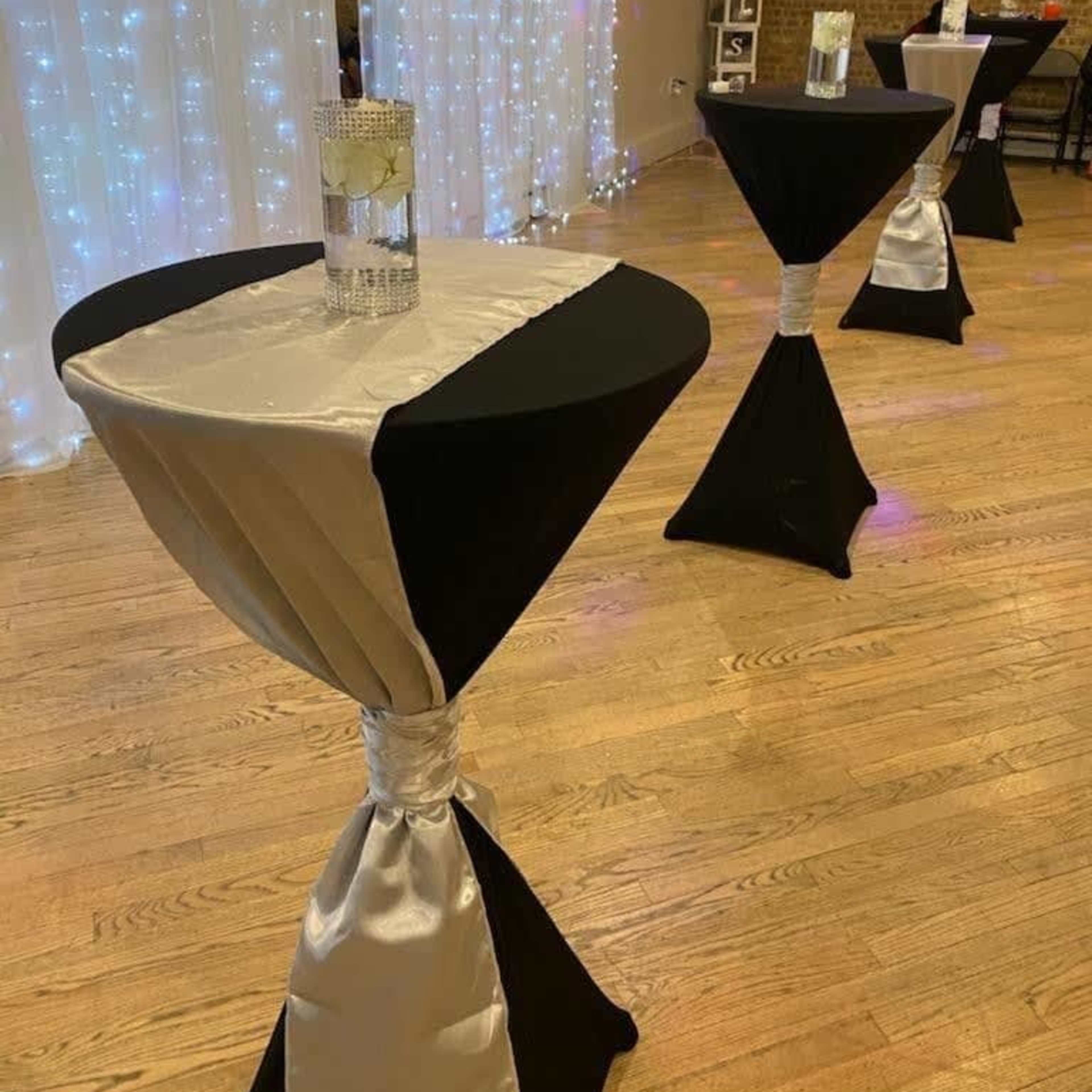 Several black and silver tablecloth-covered cocktail tables arranged in a space with fairy lights in the background.