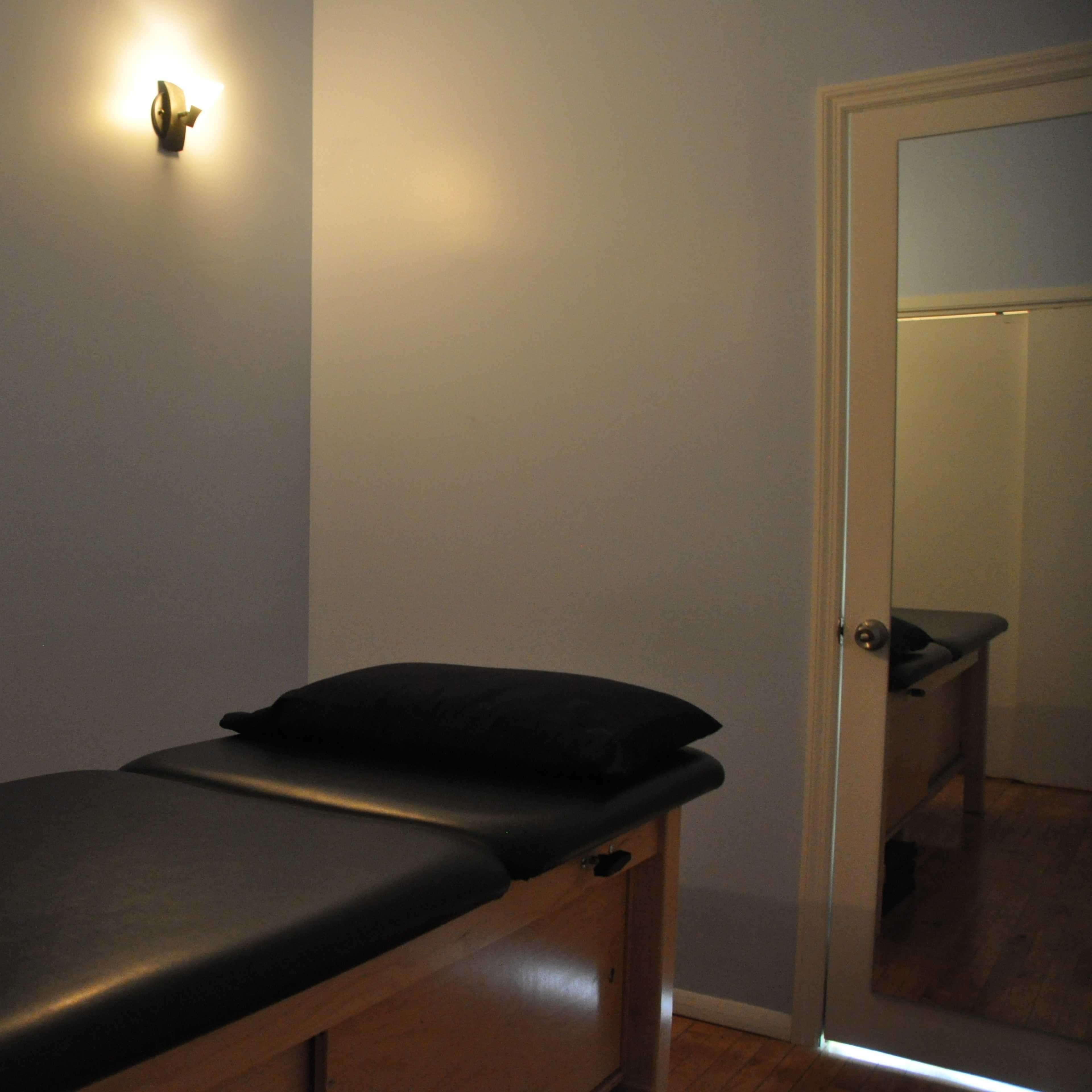 A small treatment room with a massage table, a black pillow on the table, and a mirror reflecting the room's interior.
