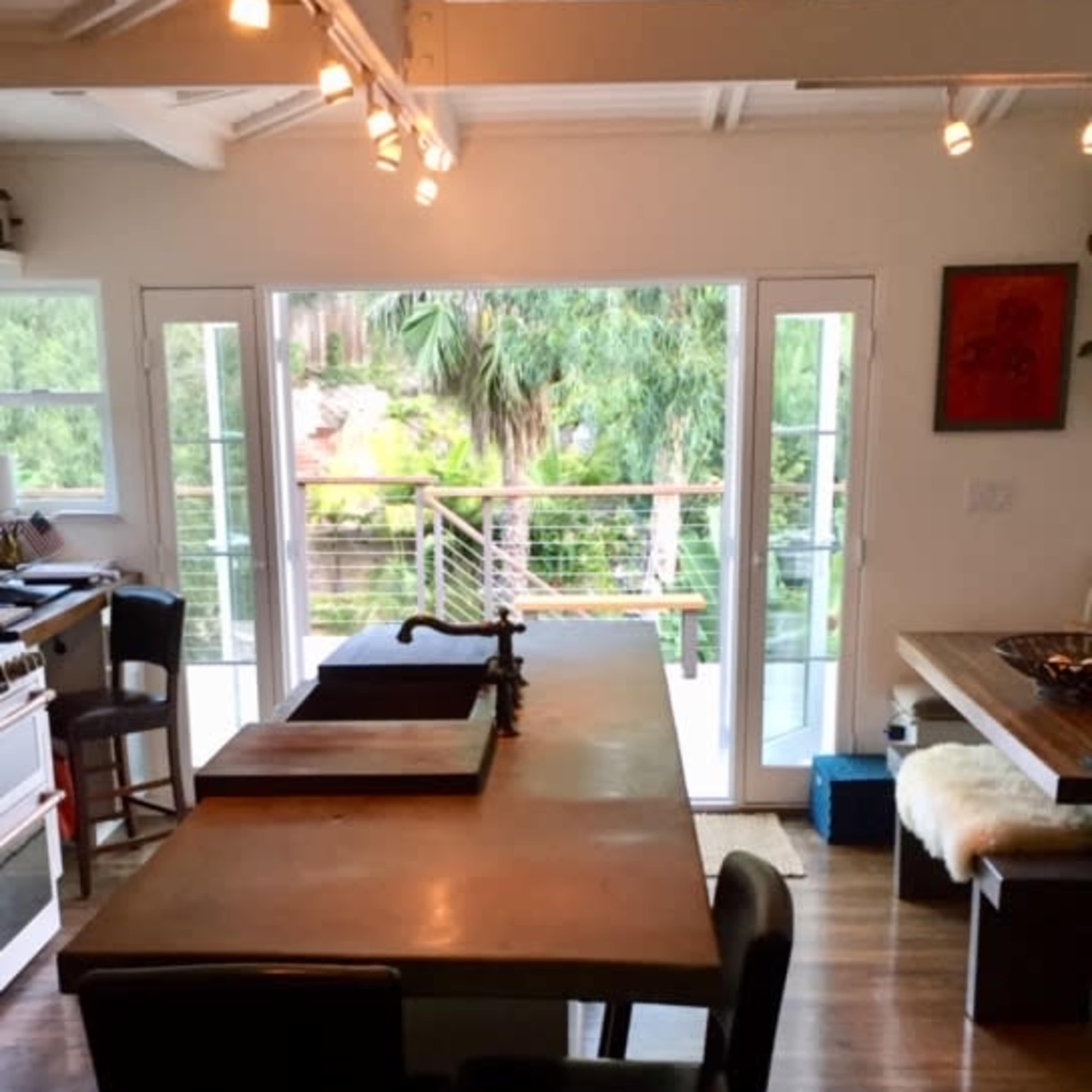A modern kitchen features an island with a sink, large windows overlooking a patio with palm trees, and sliding glass doors leading outside.