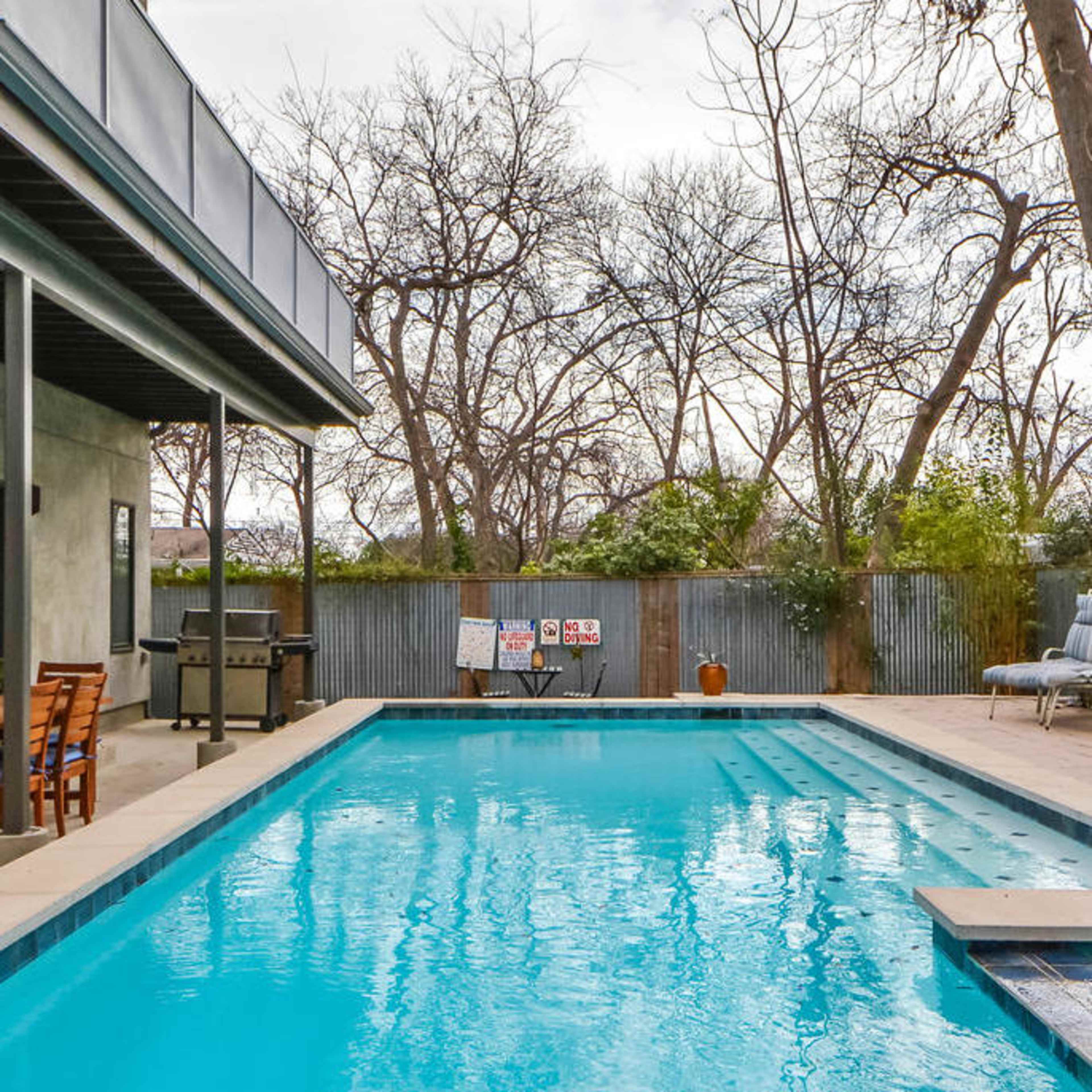 A rectangular swimming pool surrounded by a concrete patio, with a backdrop of bare trees and a corrugated metal fence.