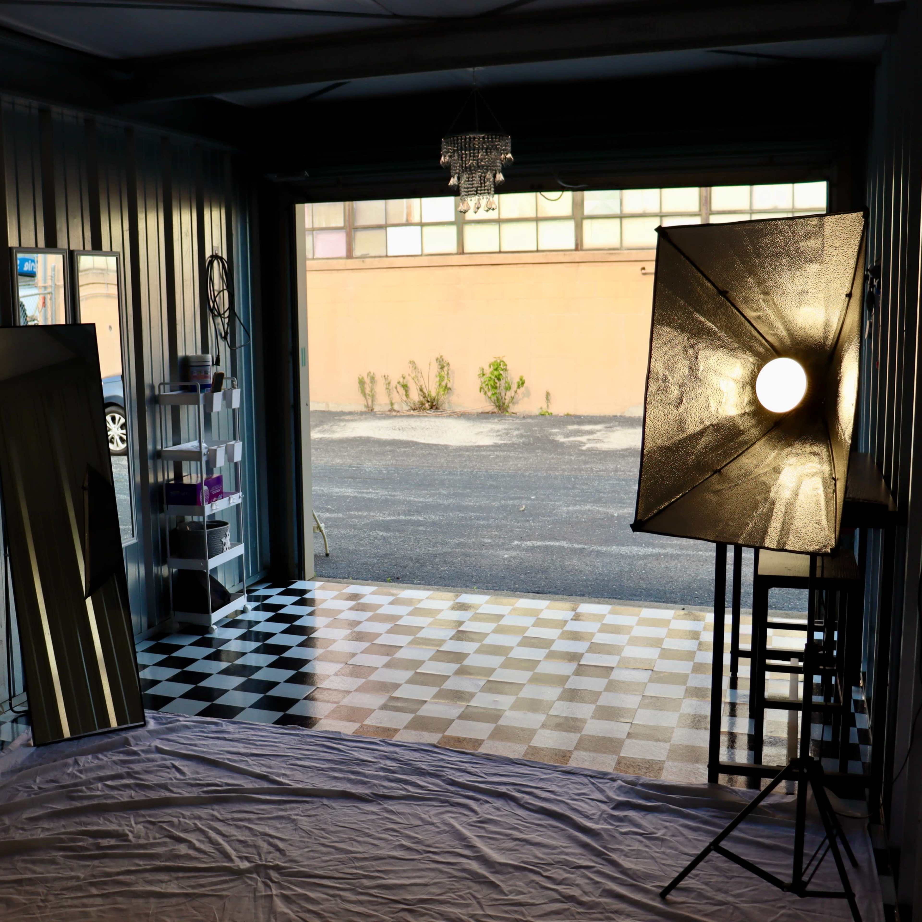 A garage studio set up with two large softbox lights, a mirror, and a checkered floor leading to an open doorway.