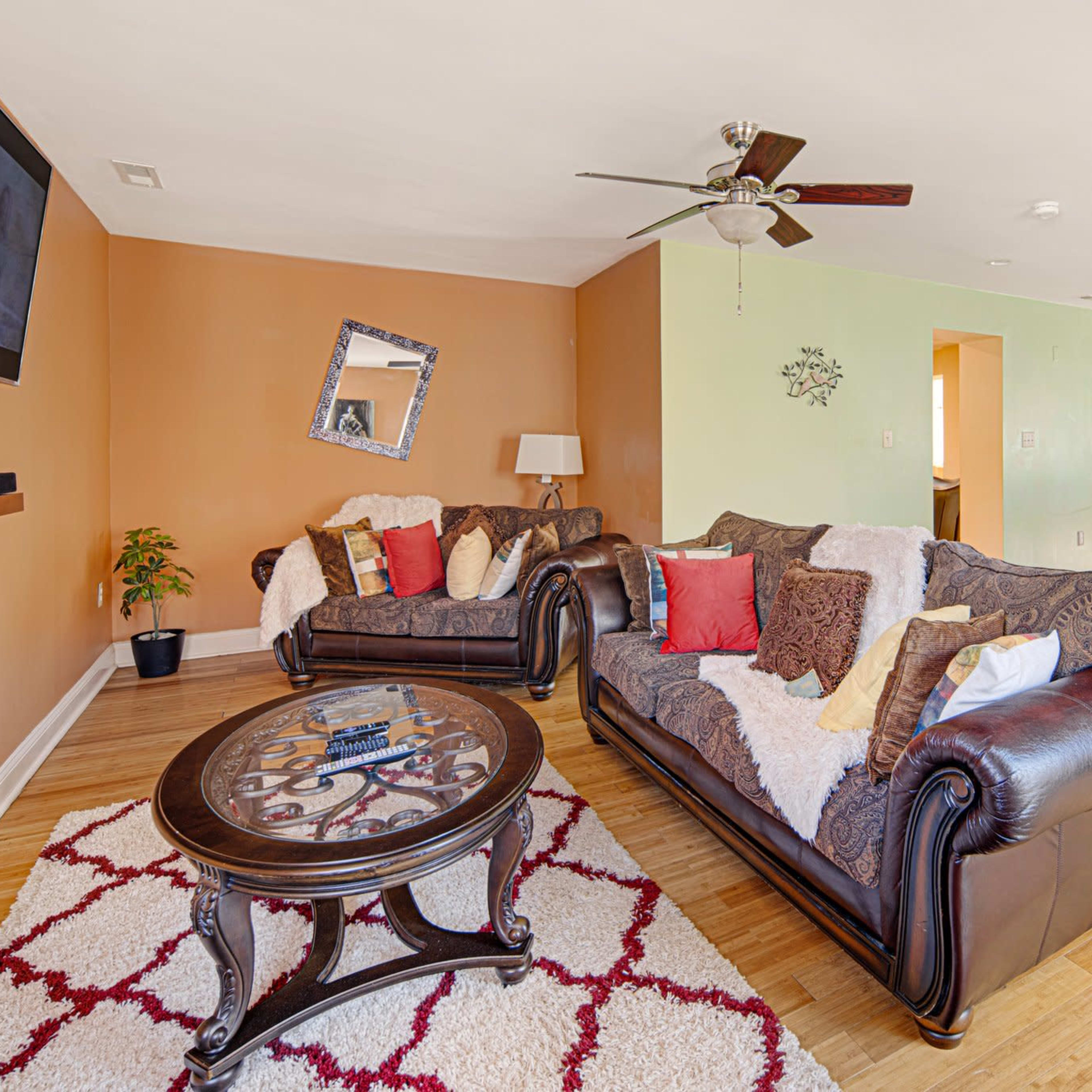 The living room features two dark leather sofas with decorative pillows arranged around a round coffee table on a patterned rug, with an open kitchen visible in the background.