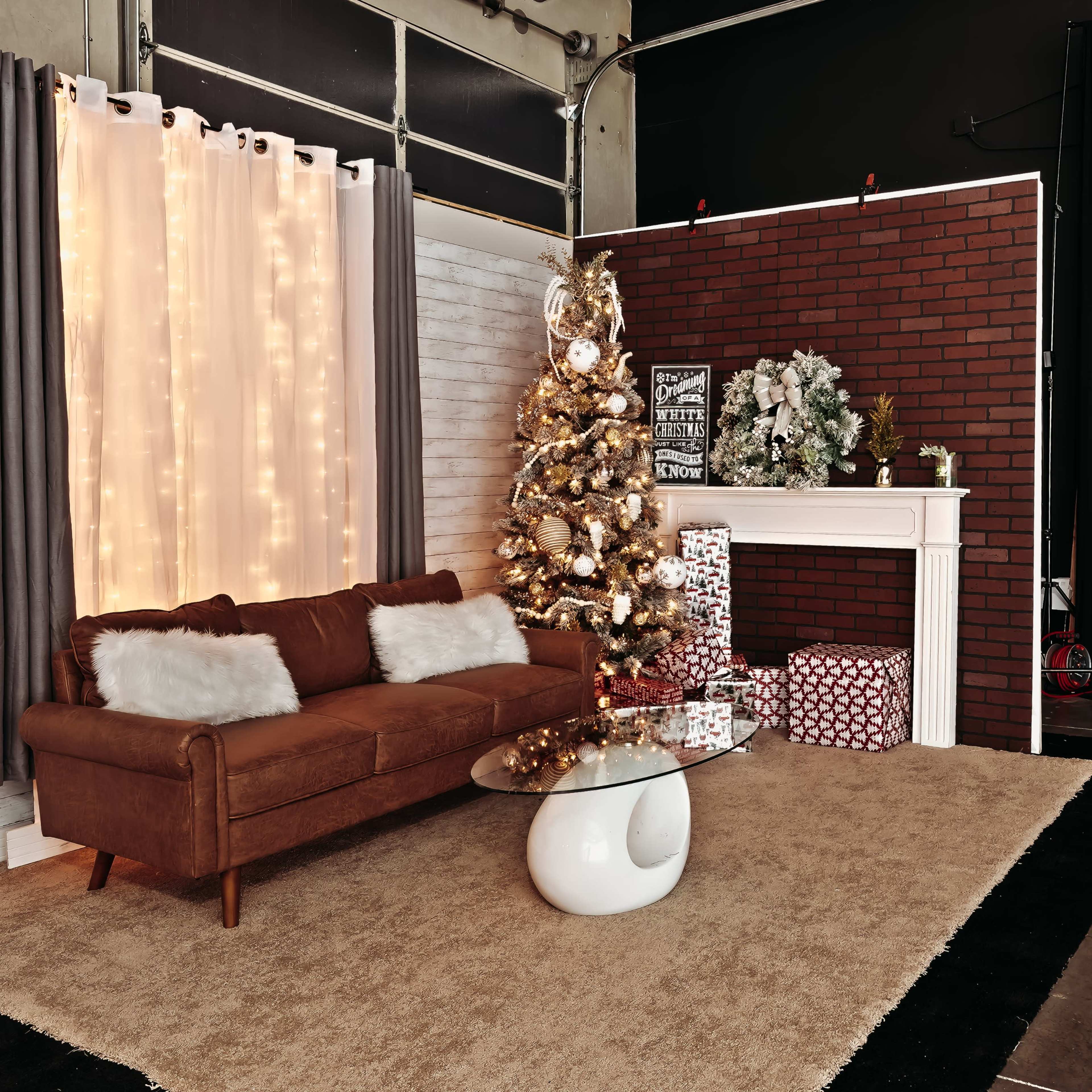A cozy room setup featuring a brown sofa, a glass coffee table, a decorated Christmas tree, and a white fireplace with presents underneath.
