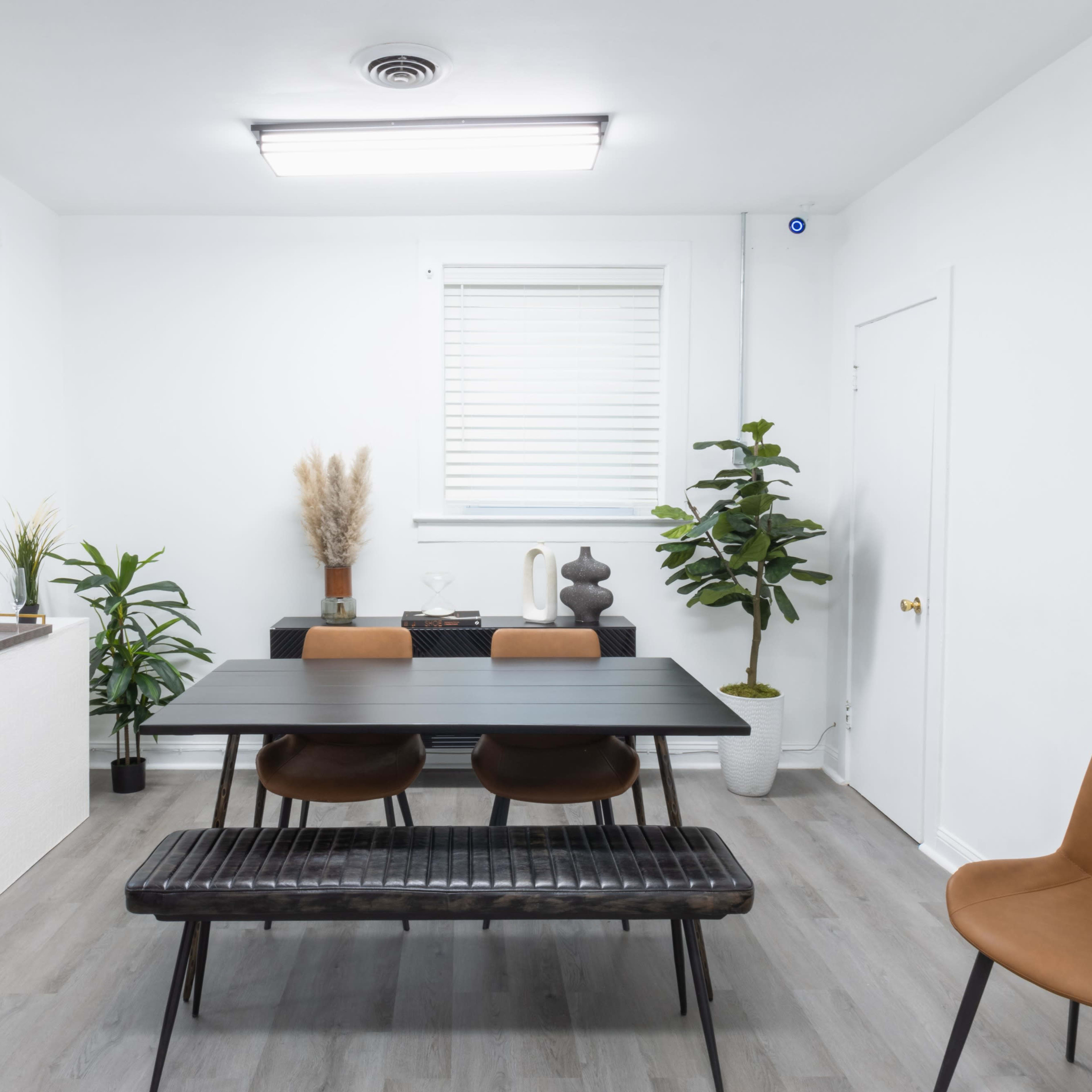 A minimalist dining room features a black table with four chairs, a bench, and decorative plants against white walls.