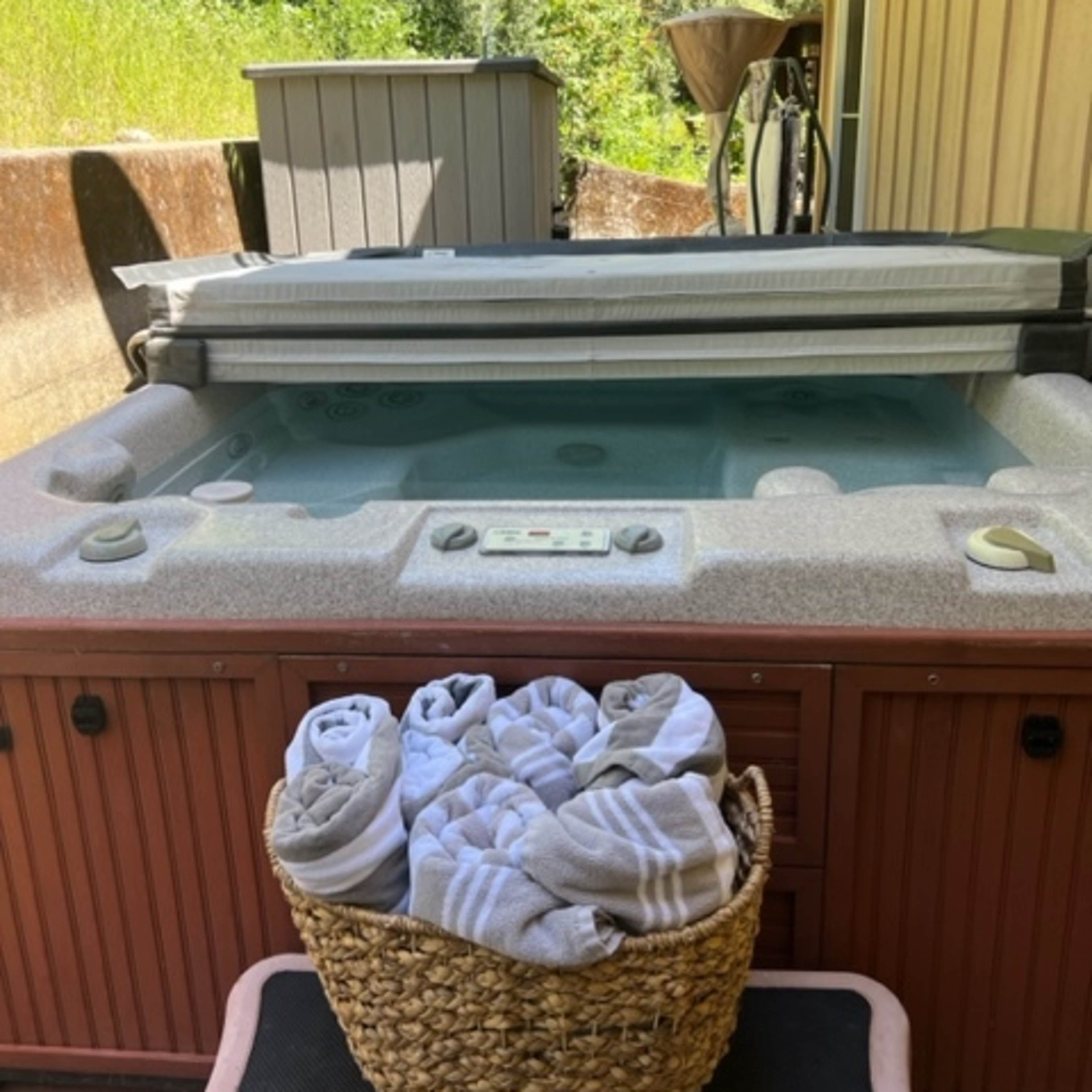 A hot tub with a cover and a basket of towels sits beside a wooden deck and a shed, surrounded by greenery.