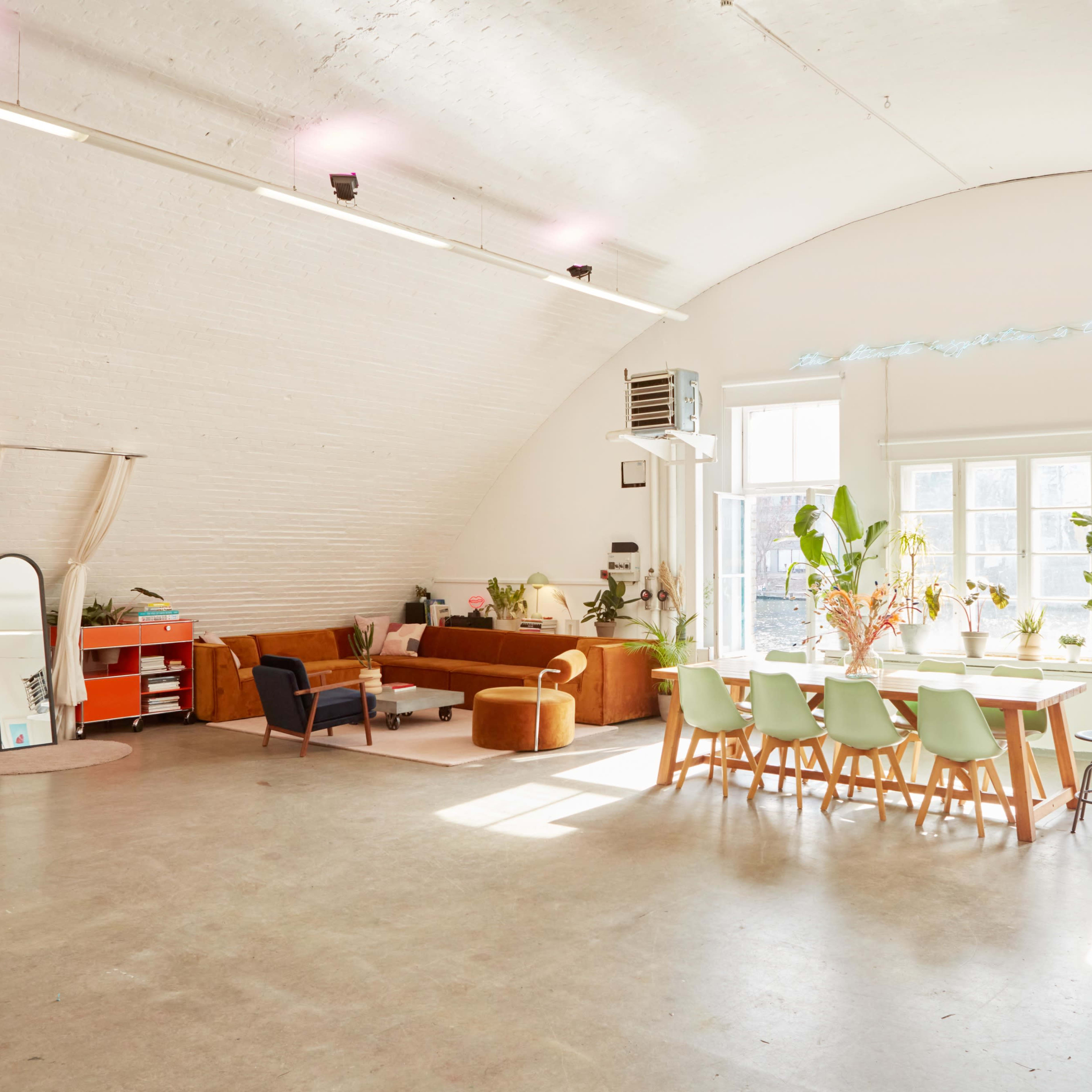 A spacious, well-lit room with a curved ceiling, featuring a seating area with a sofa, a dining table, and large windows filled with plants.