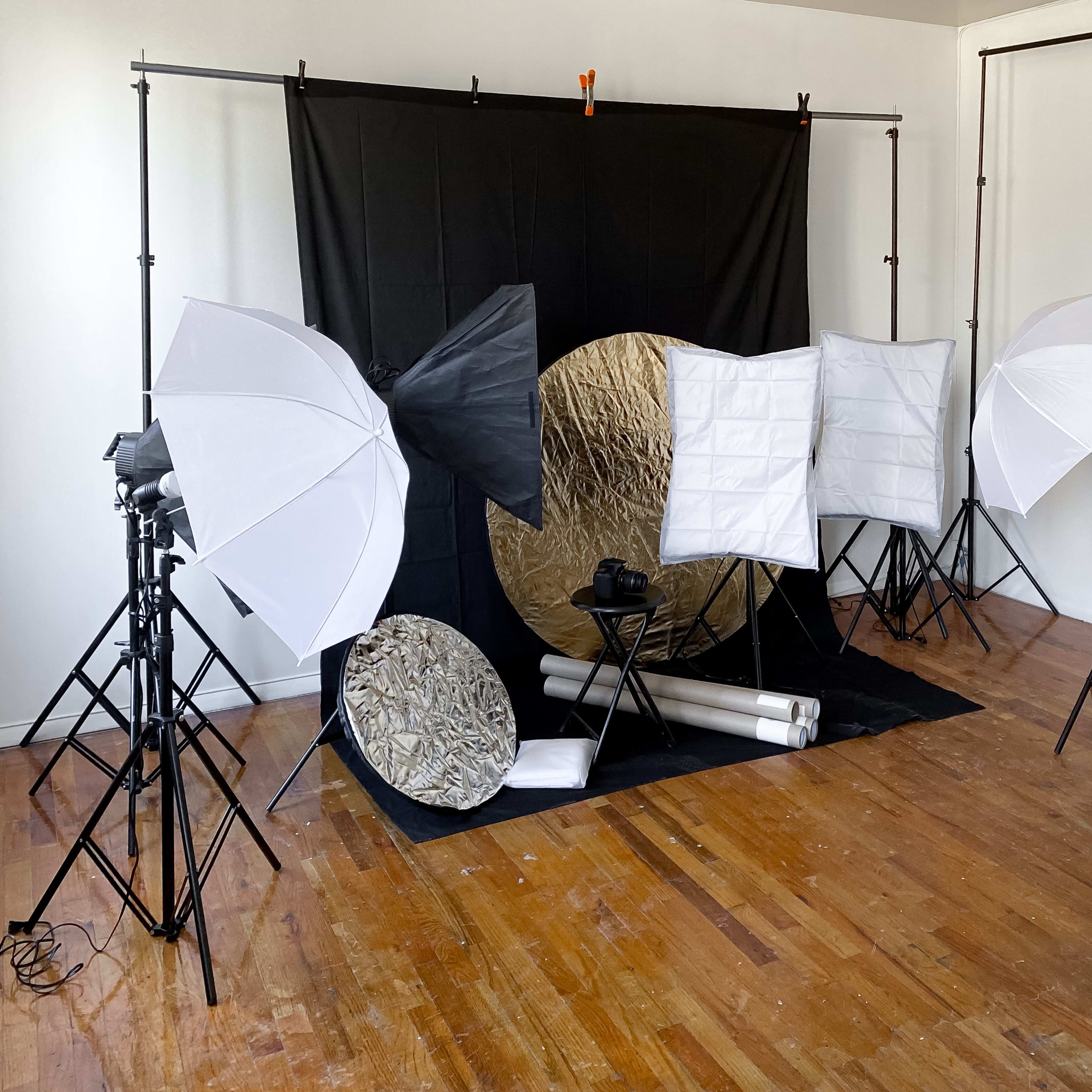 A photo studio setup featuring a black backdrop, multiple light stands with softboxes, and reflector discs arranged on a wooden floor.