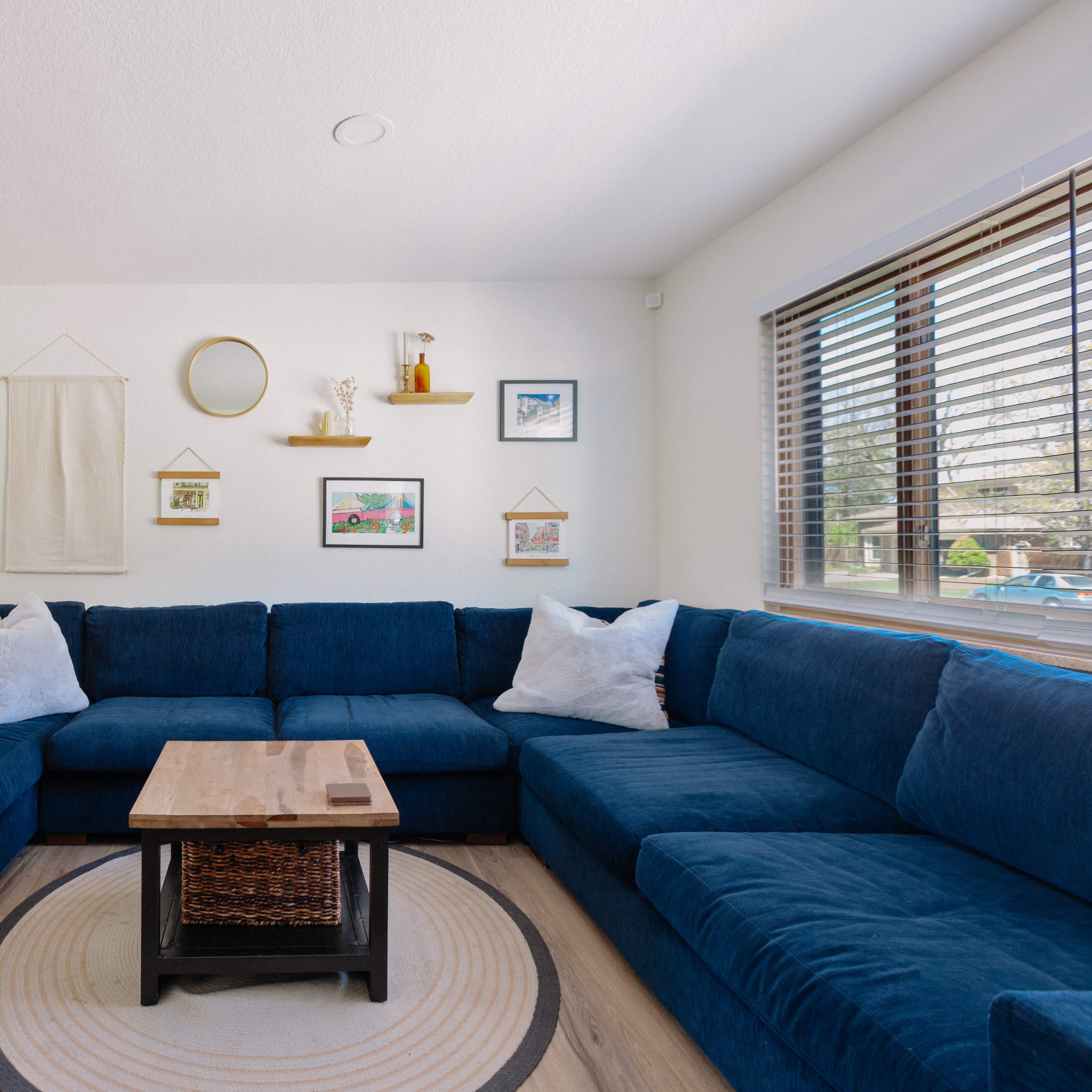 A spacious living room features a large blue sectional sofa, a wooden coffee table, and framed artwork on the walls beside a window with blinds.