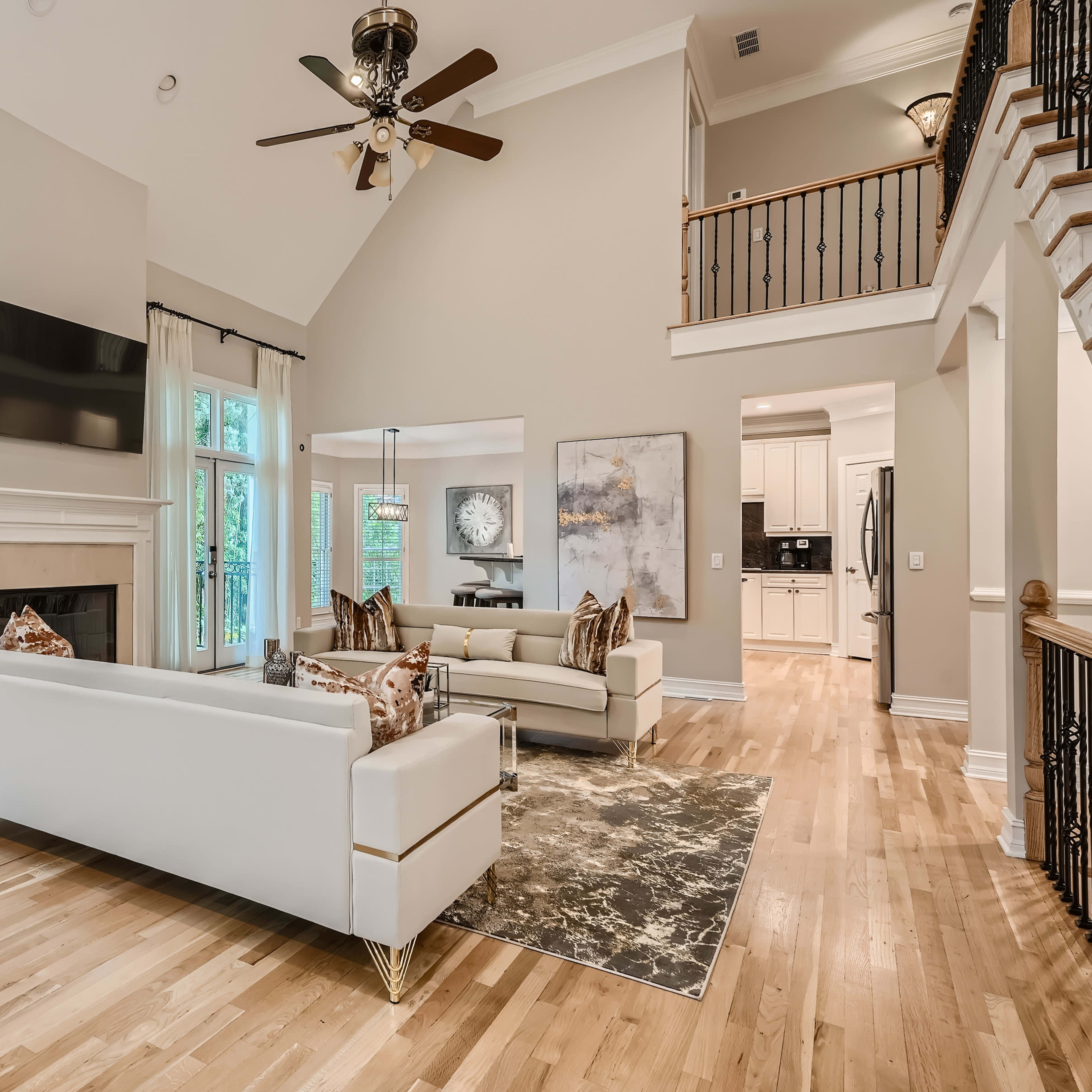 A spacious living room with white sofas, a large flat-screen TV mounted on the wall, and hardwood flooring, illuminated by natural light from large windows.