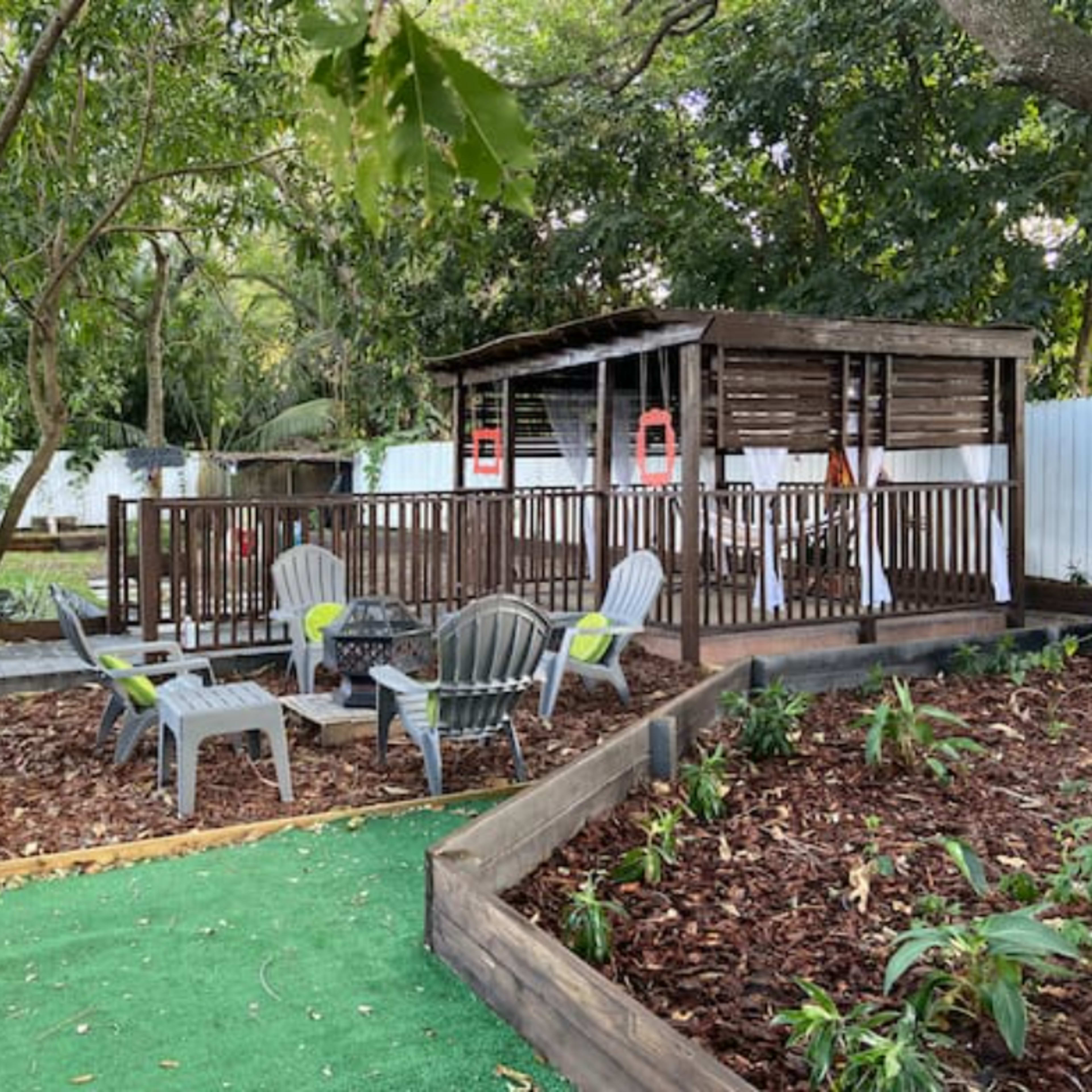 A backyard with a wooden deck featuring seating areas, a garden with mulch, and a shaded gazebo surrounded by trees.
