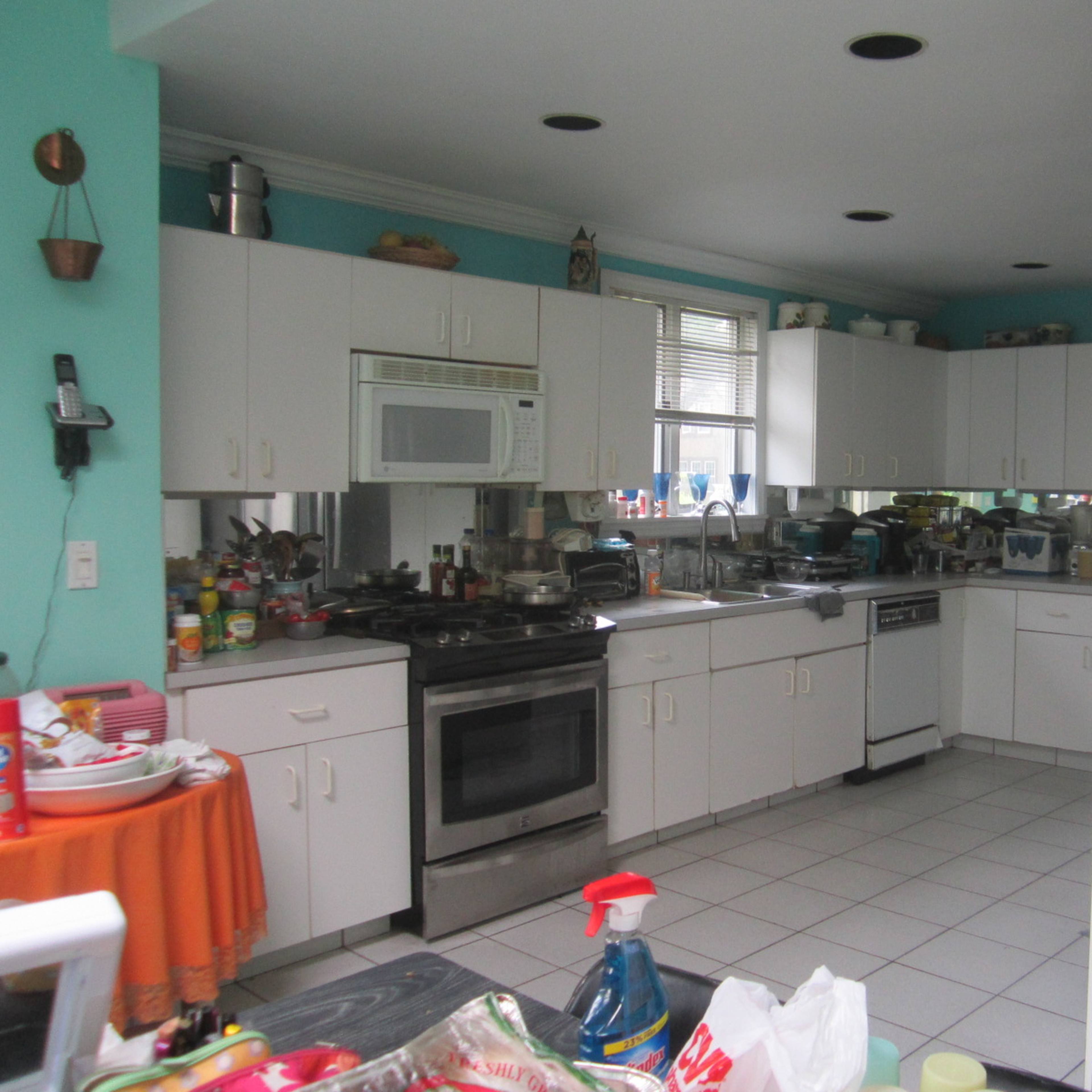A cluttered kitchen with white cabinetry, a stove, and various kitchen appliances and items scattered on the countertops.
