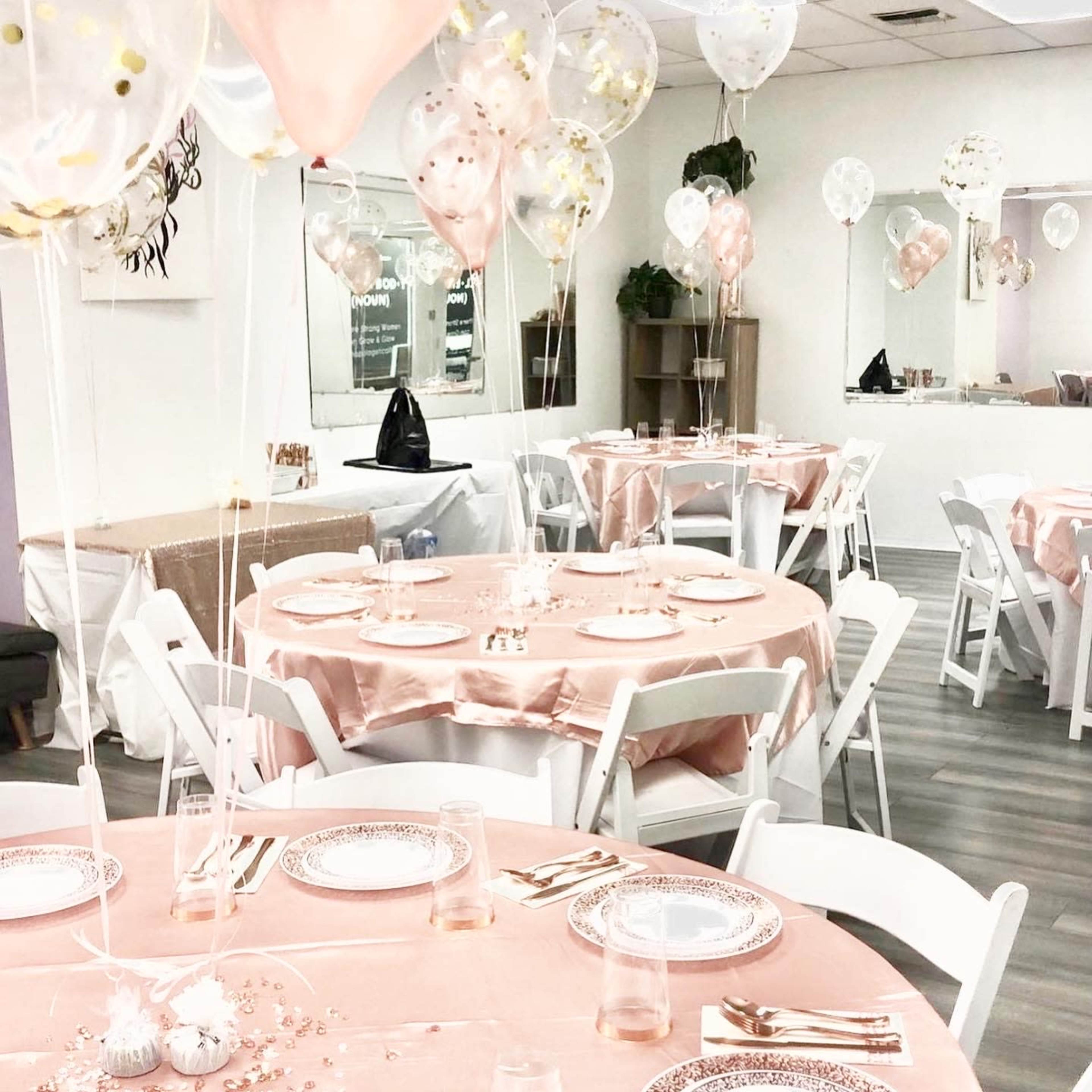 A decorated event space with round tables draped in pink tablecloths, each set with plates, glasses, and balloons.