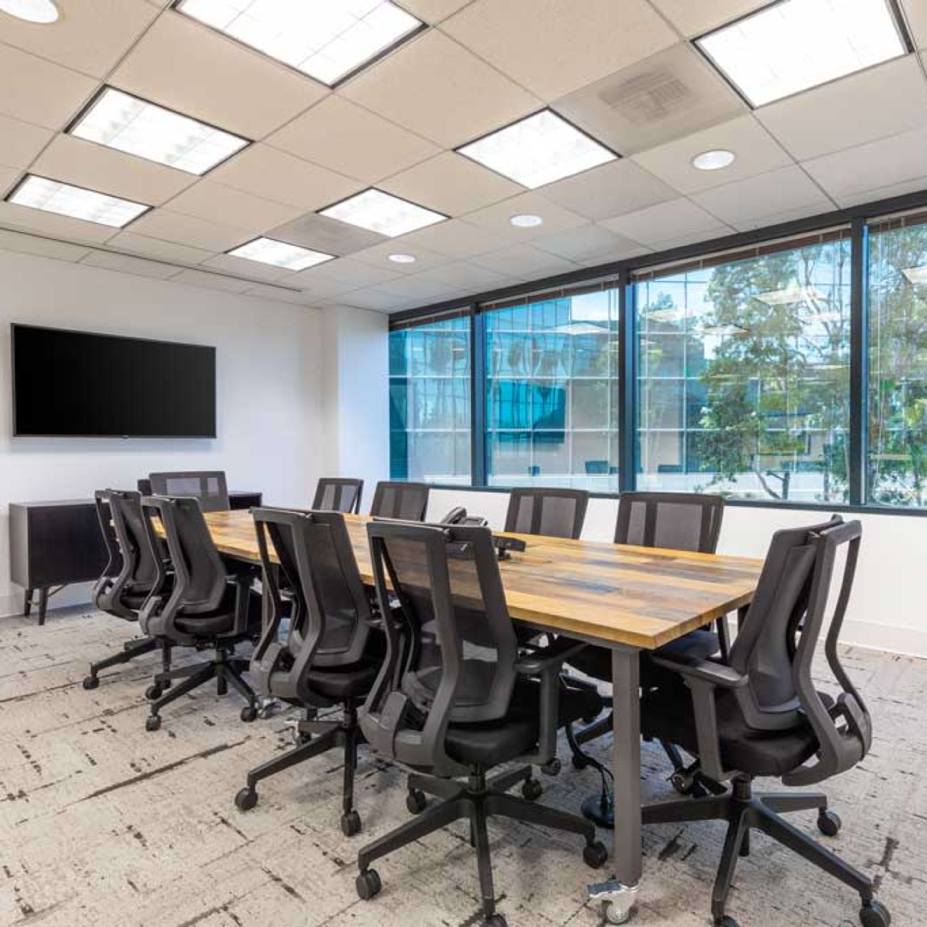 A modern meeting room features a large wooden table surrounded by ergonomic chairs, with large windows providing an outdoor view and a wall-mounted screen.