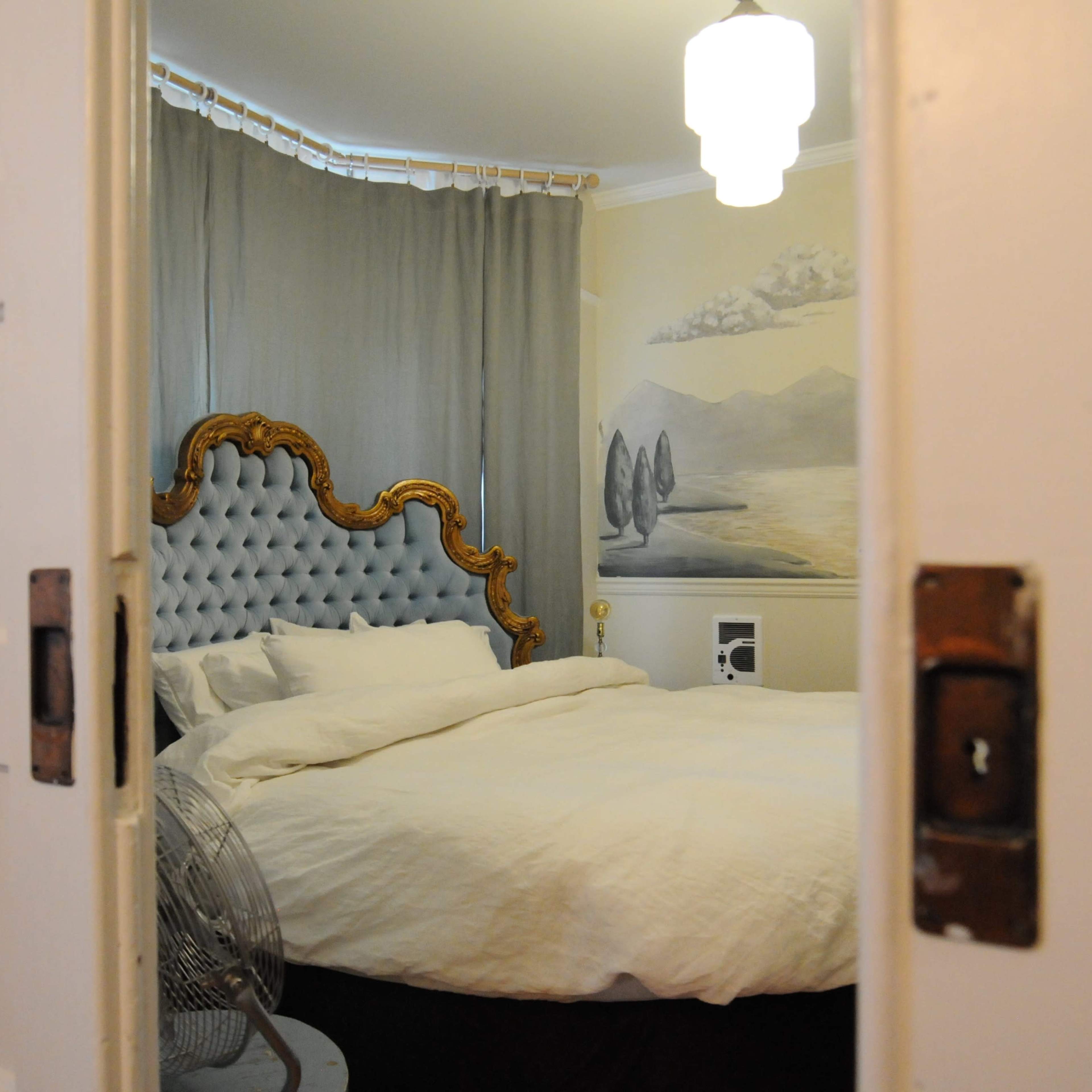 A neatly made bed with a tufted headboard is visible through an open bedroom door, framed by a light-colored wall and a decorative curtain.