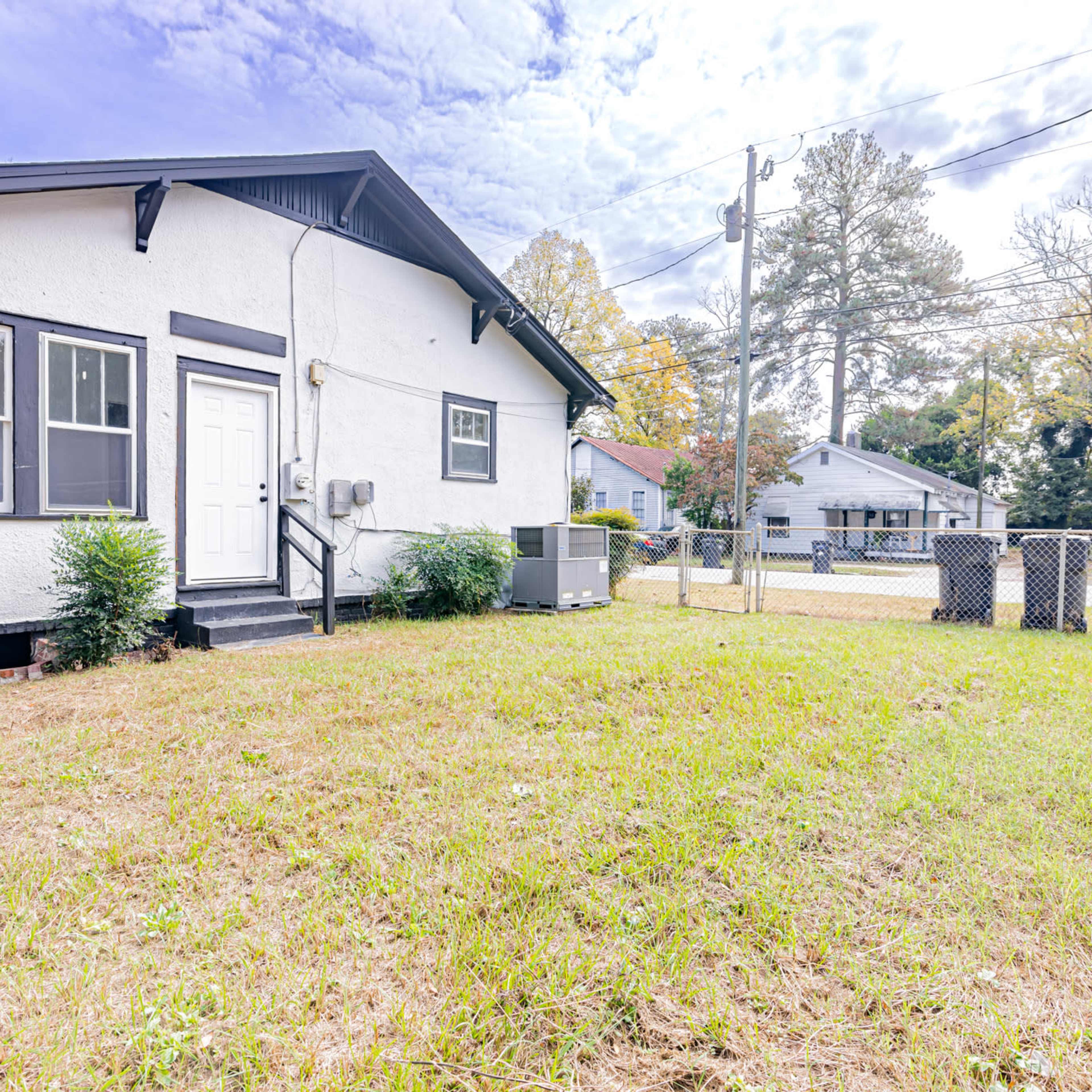 A single-story house with a white exterior, a front door, and a grassy yard, surrounded by trees and another house in the background.