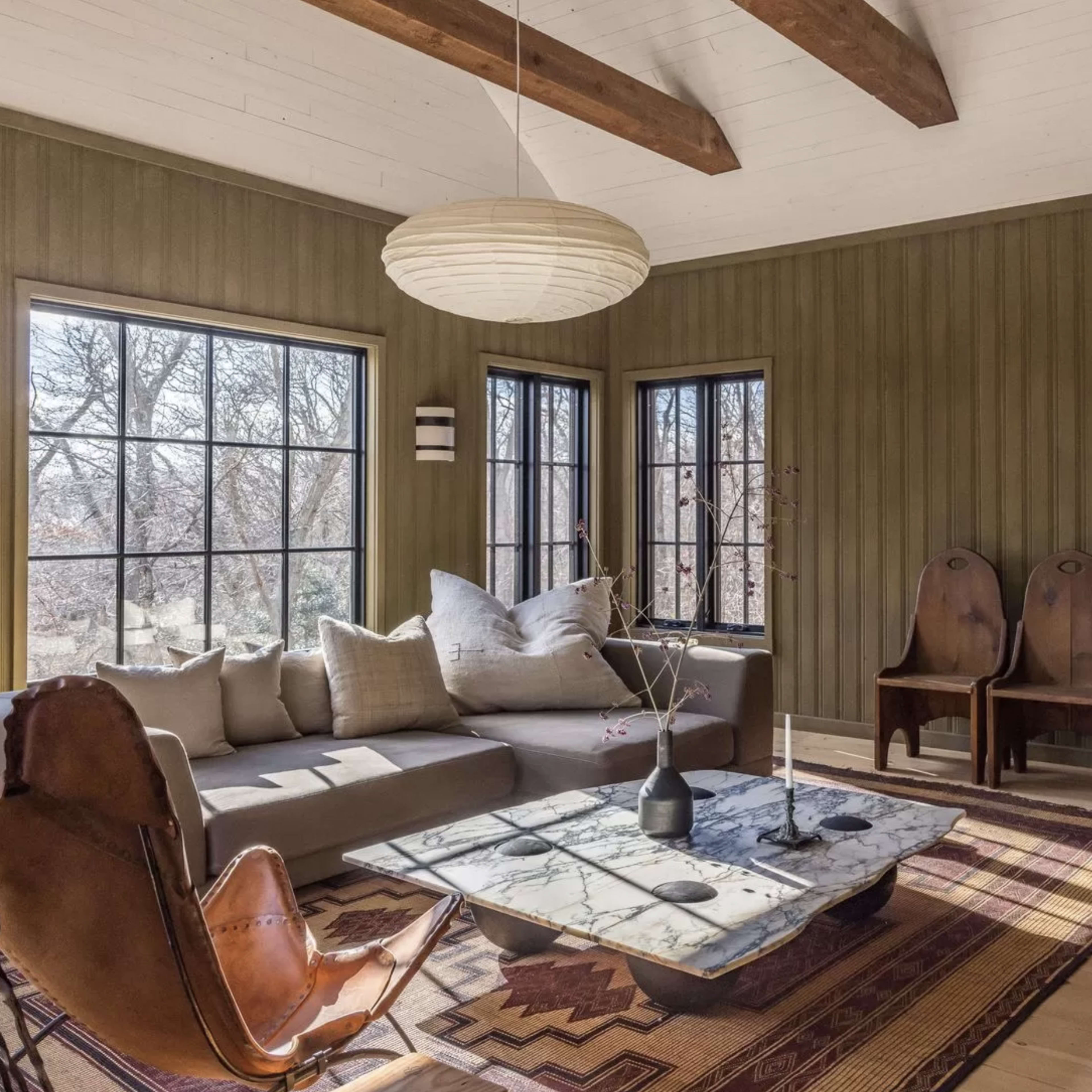 A cozy living room with a sectional sofa, a coffee table, a patterned rug, and two wooden chairs against a wall of windows.