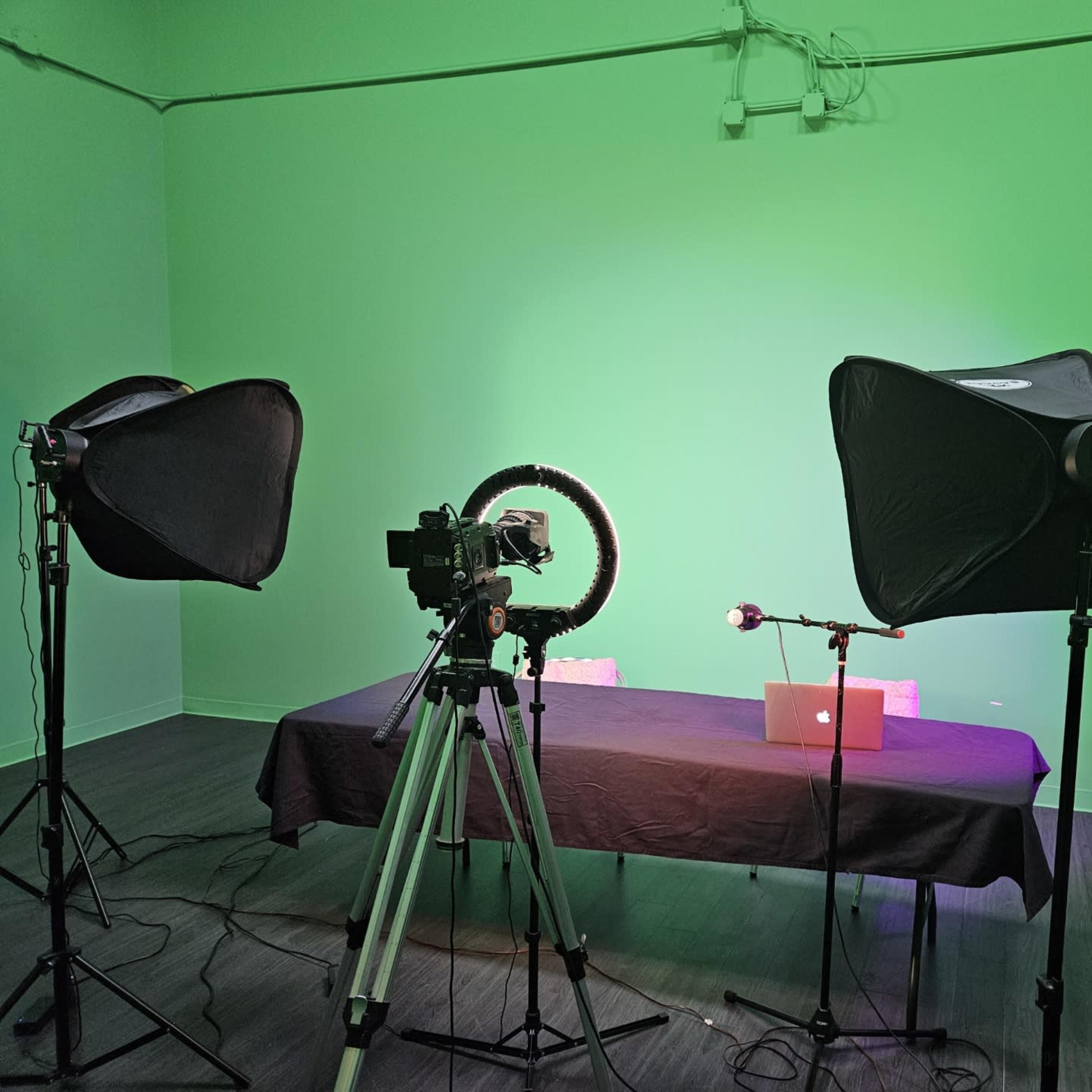 A video recording setup featuring a table with a laptop, a camera on a tripod, softbox lights, and a ring light in front of a green background.