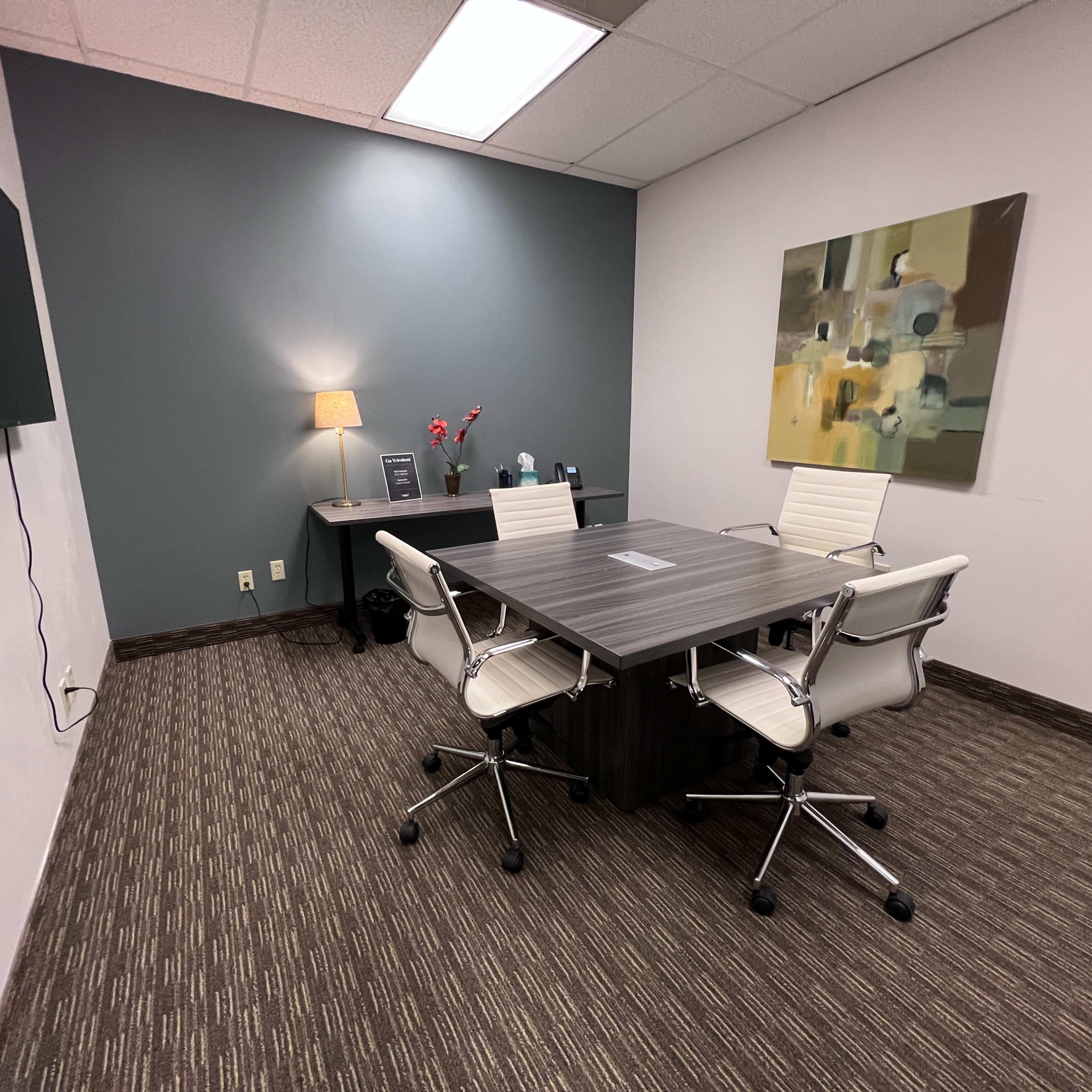 A small conference room with a rectangular table, two white chairs, a desk with a lamp, and an abstract painting on the wall.