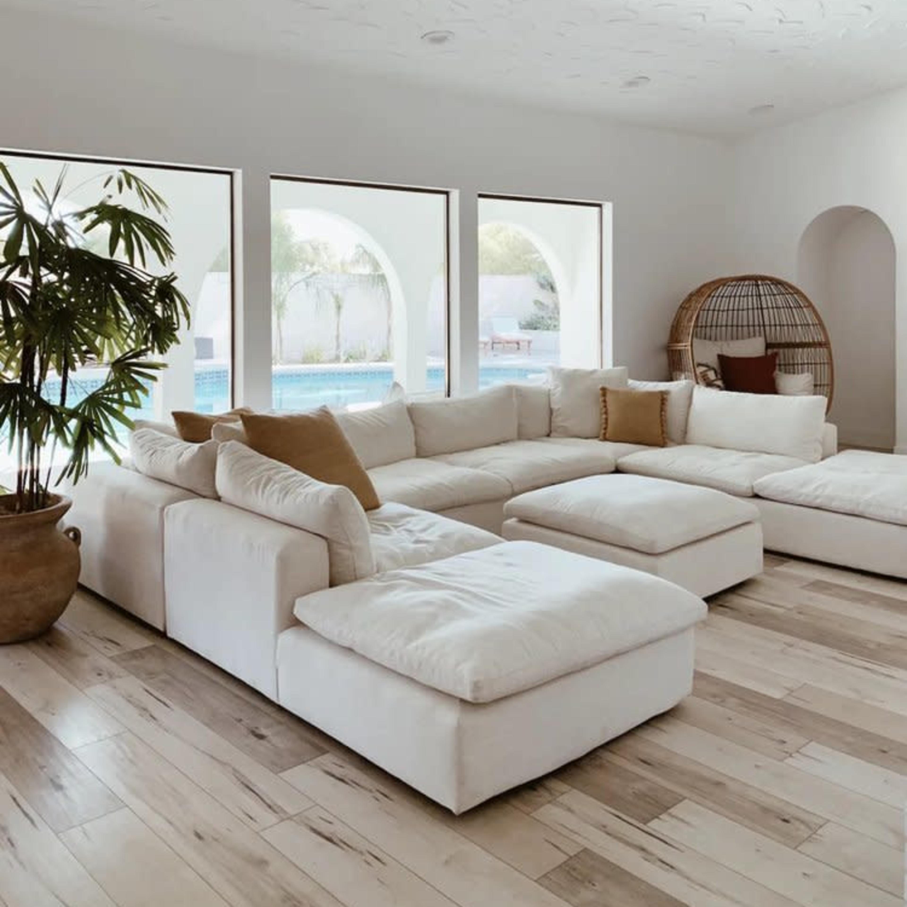 A light-filled living room with a large white sectional sofa, a decorative plant, and arched windows revealing a pool view.
