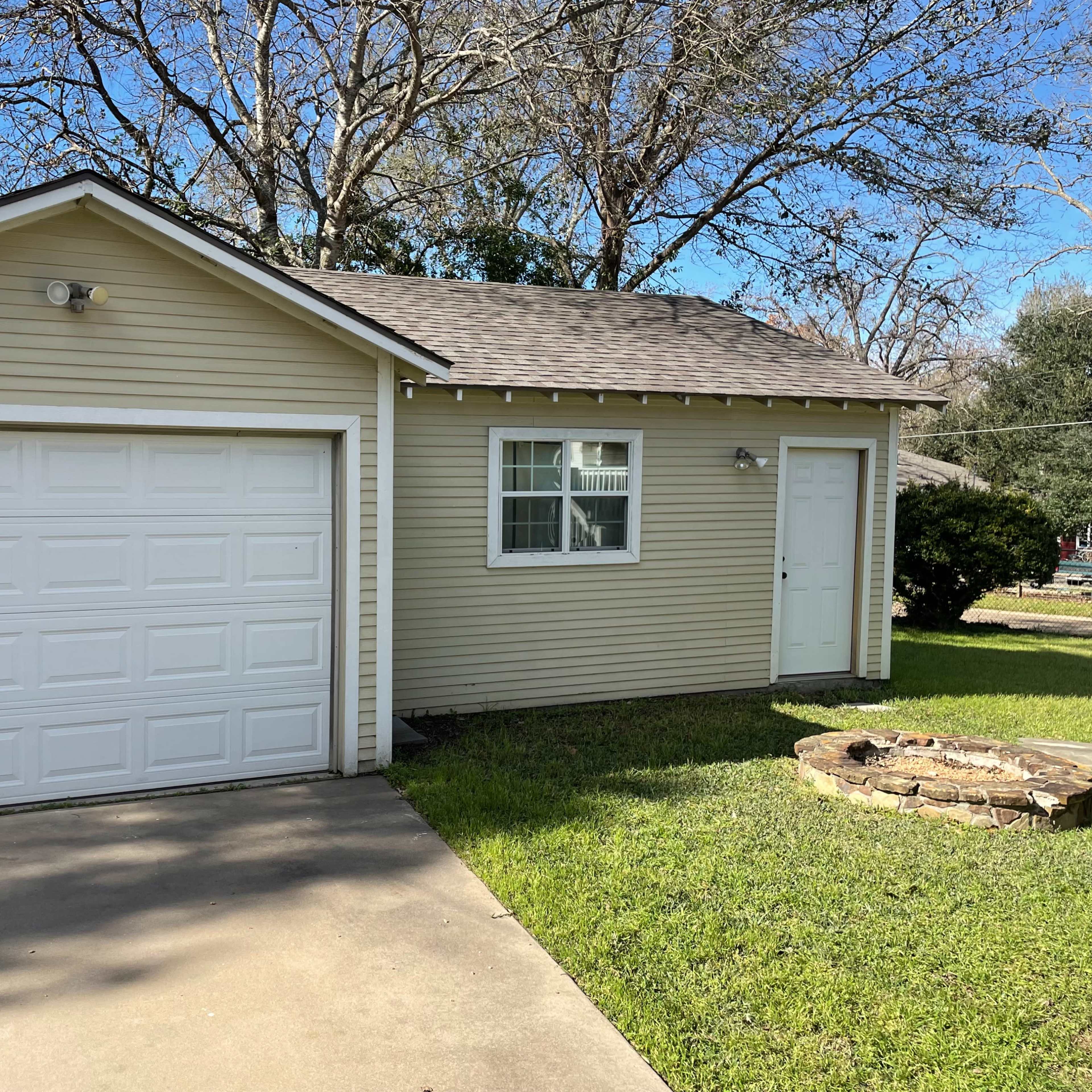 A single-story house with a beige exterior, a white garage door, and a small stone fire pit in the front yard.