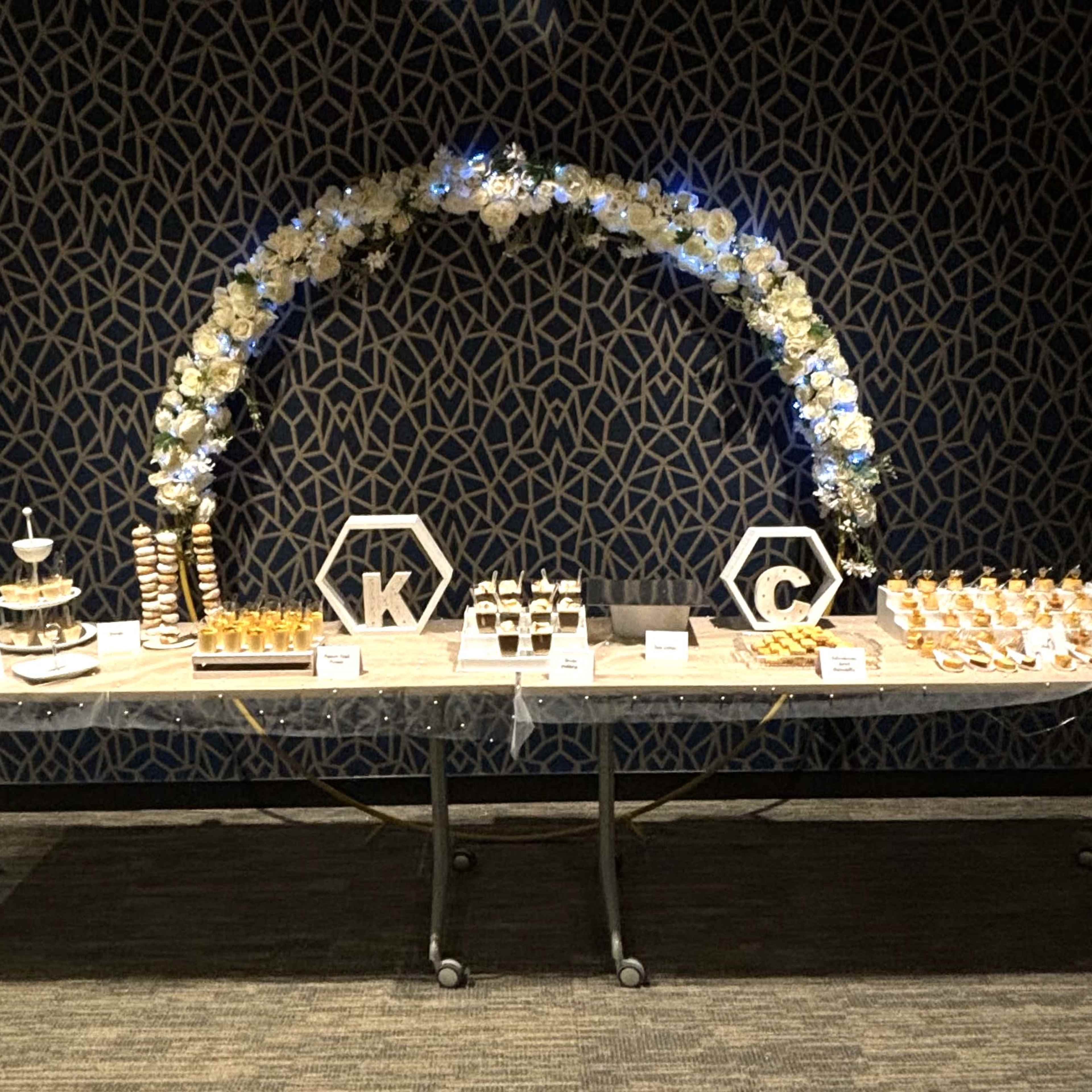 A dessert display featuring assorted pastries and a floral arch with the letters "K" and "C" arranged on a table.