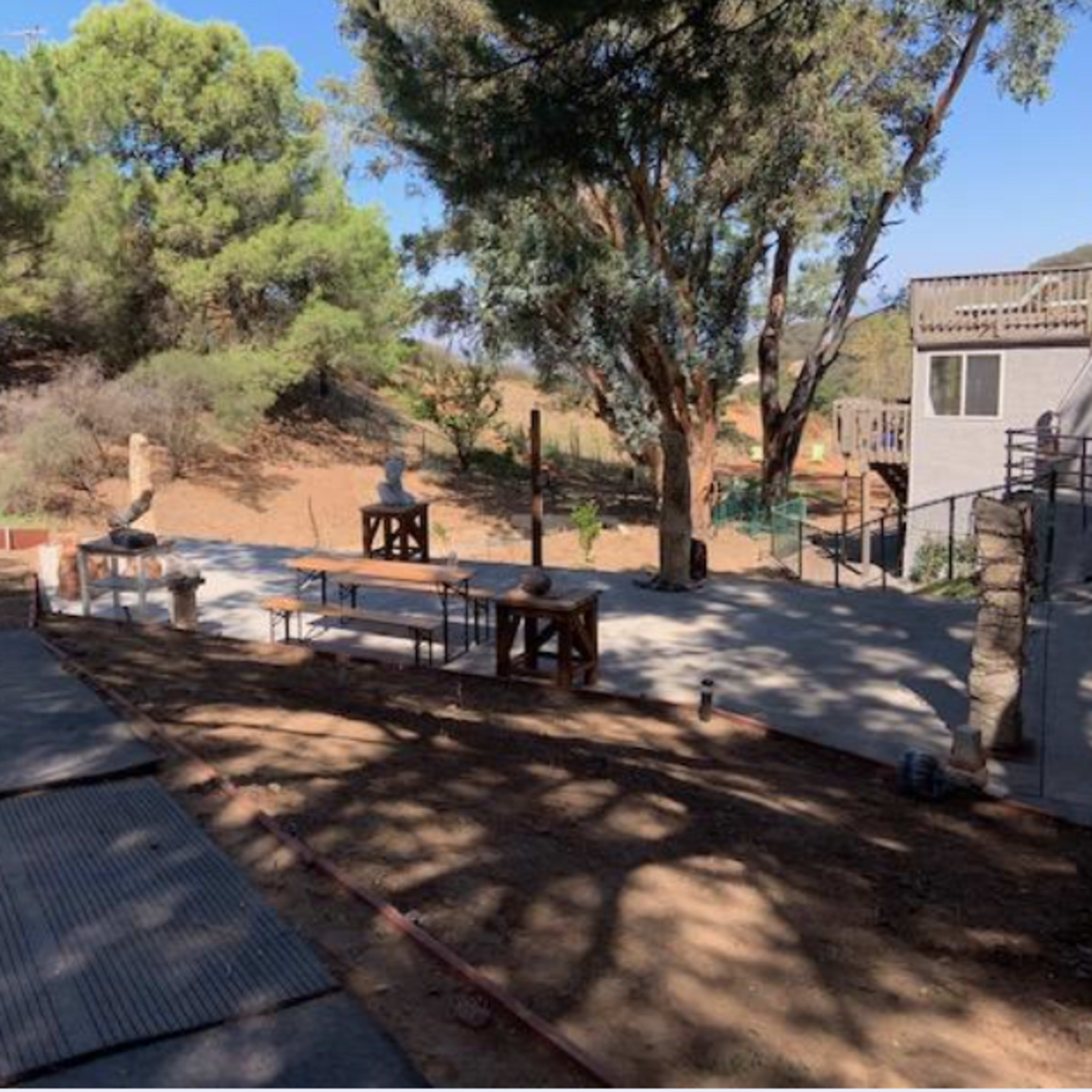 A landscaped outdoor area featuring a concrete patio with wooden tables and benches, surrounded by trees and hillside.