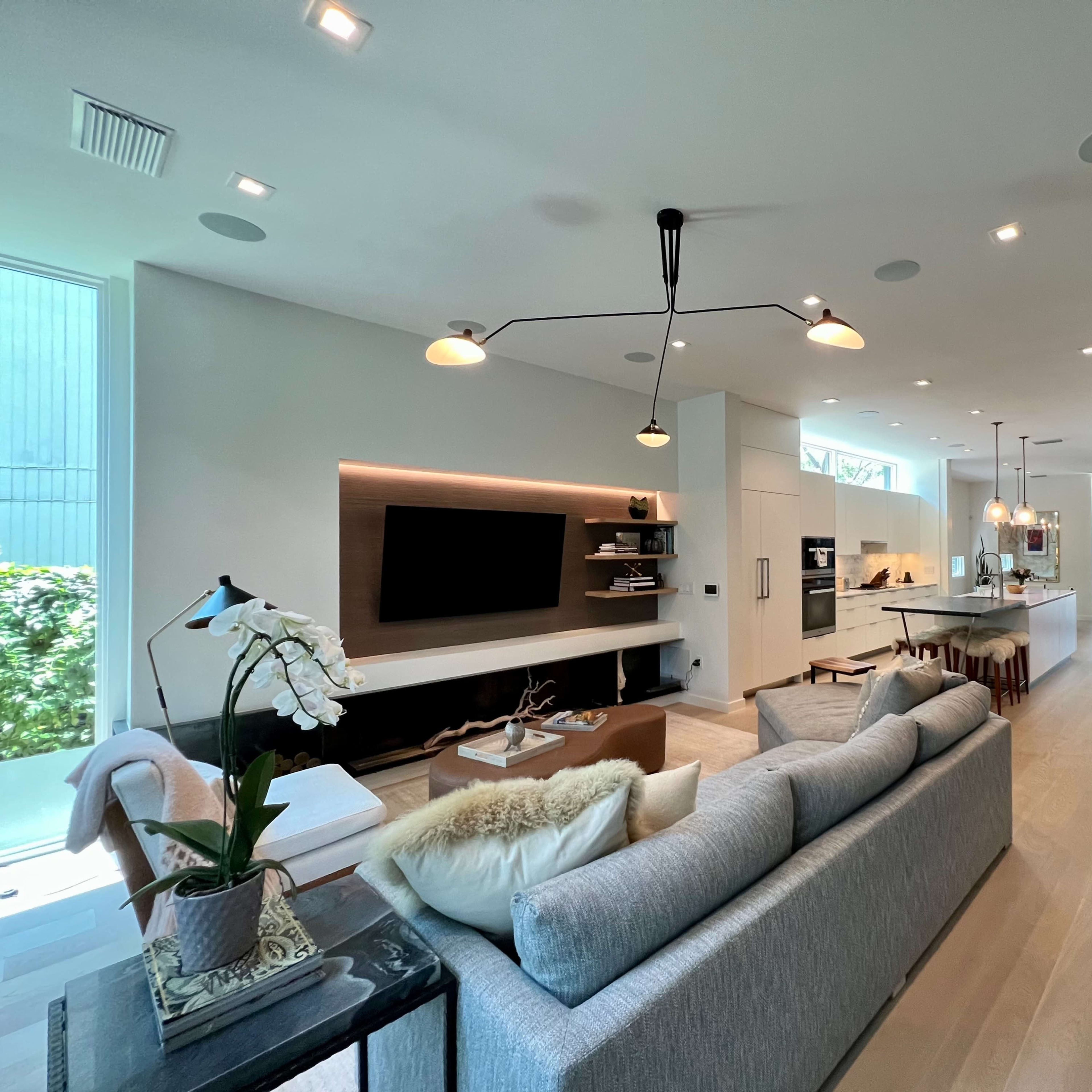 A modern living room connected to a kitchen, featuring a light gray sofa, a wall-mounted television, and large windows that allow natural light to enter the space.