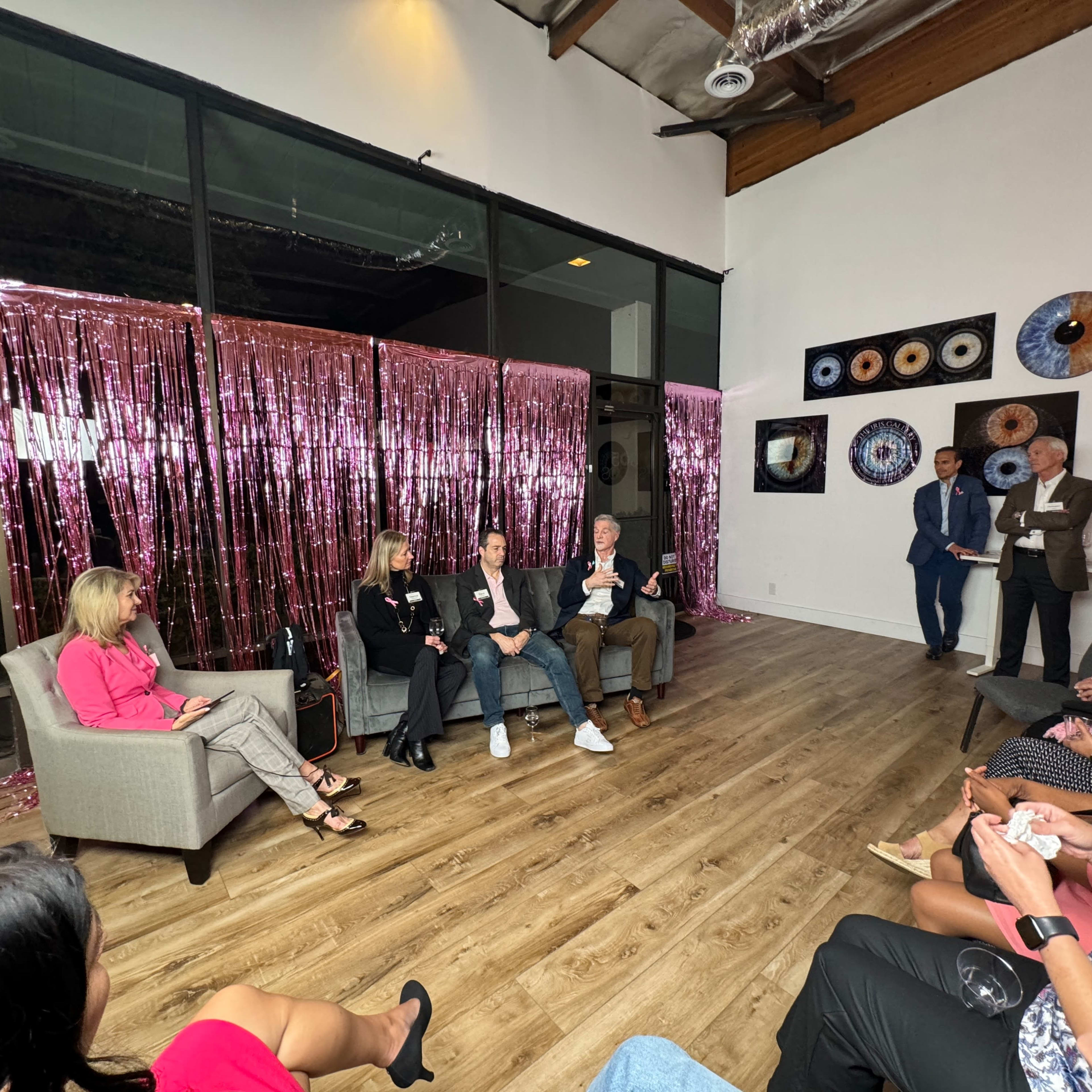 A panel discussion is taking place in a well-lit room with pink decorative curtains, featuring several participants seated on a couch while an audience listens.