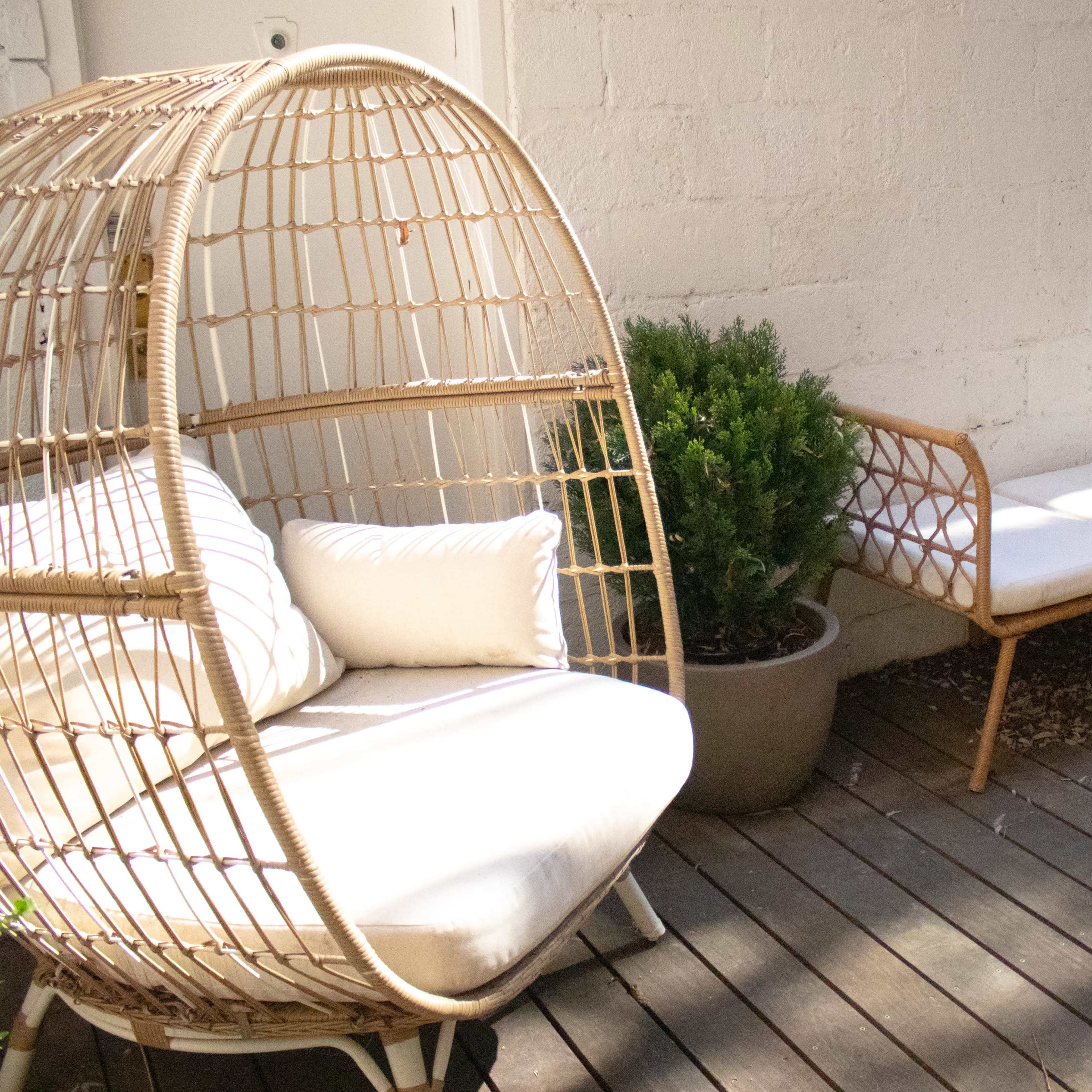 A wicker chair and a bench with cushions sit on a wooden deck next to decorative potted plants in a sunlit outdoor space.