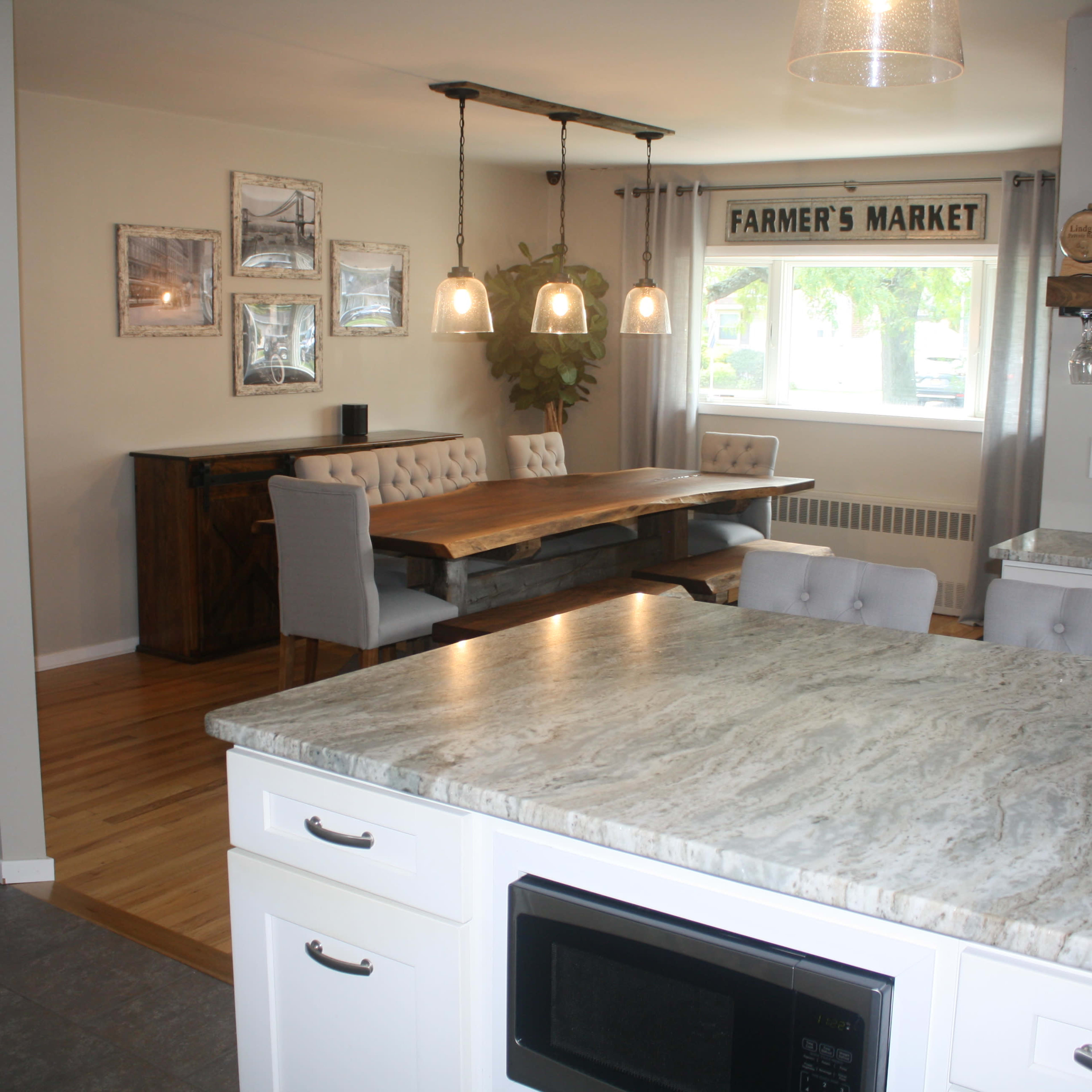 A kitchen with a marble countertop and white cabinets, overlooking a dining area featuring a wooden table and grey chairs under a sign reading "FARMER'S MARKET."