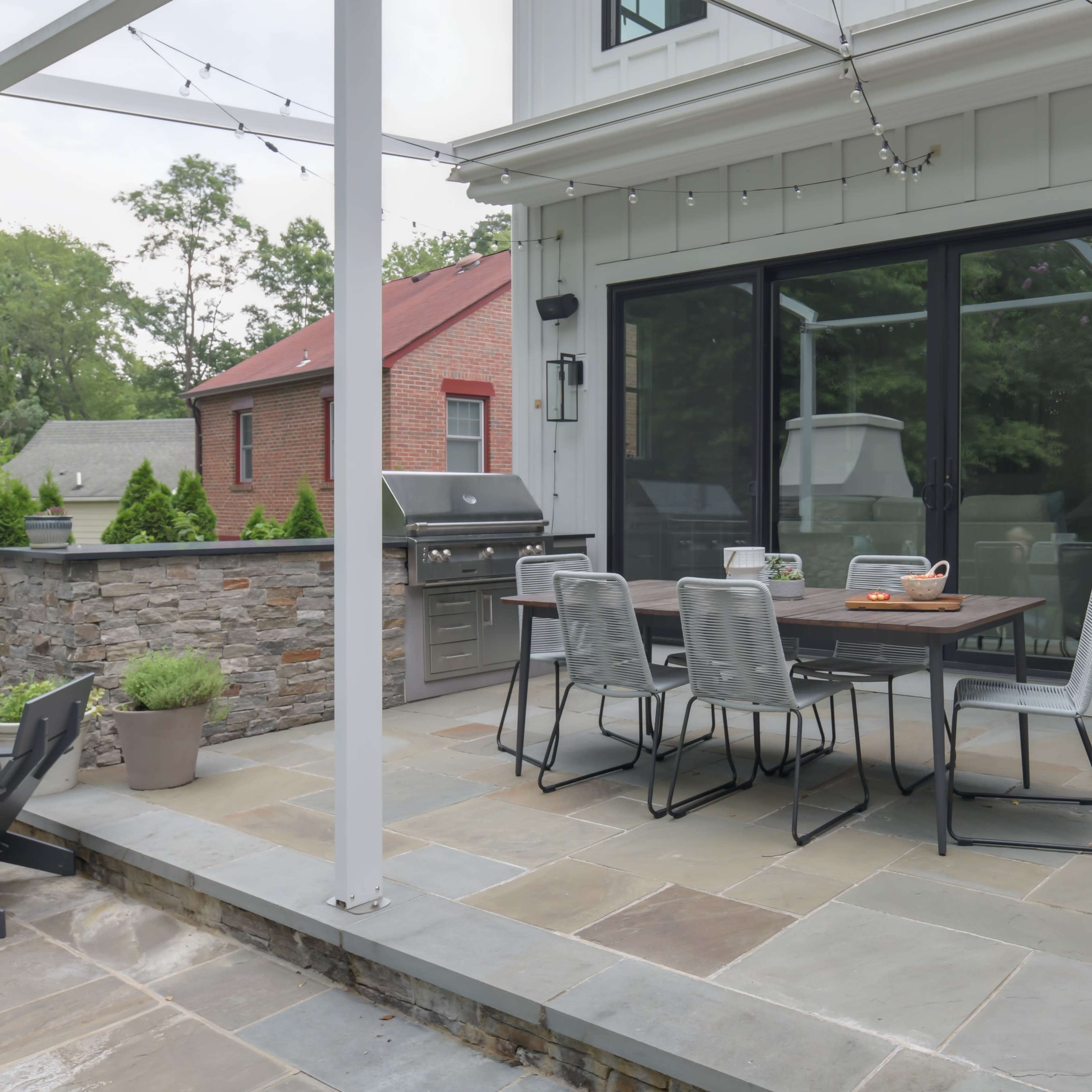 A patio area features a dining table with chairs, a barbecue grill, and string lights, surrounded by a stone wall and planters with flowers.