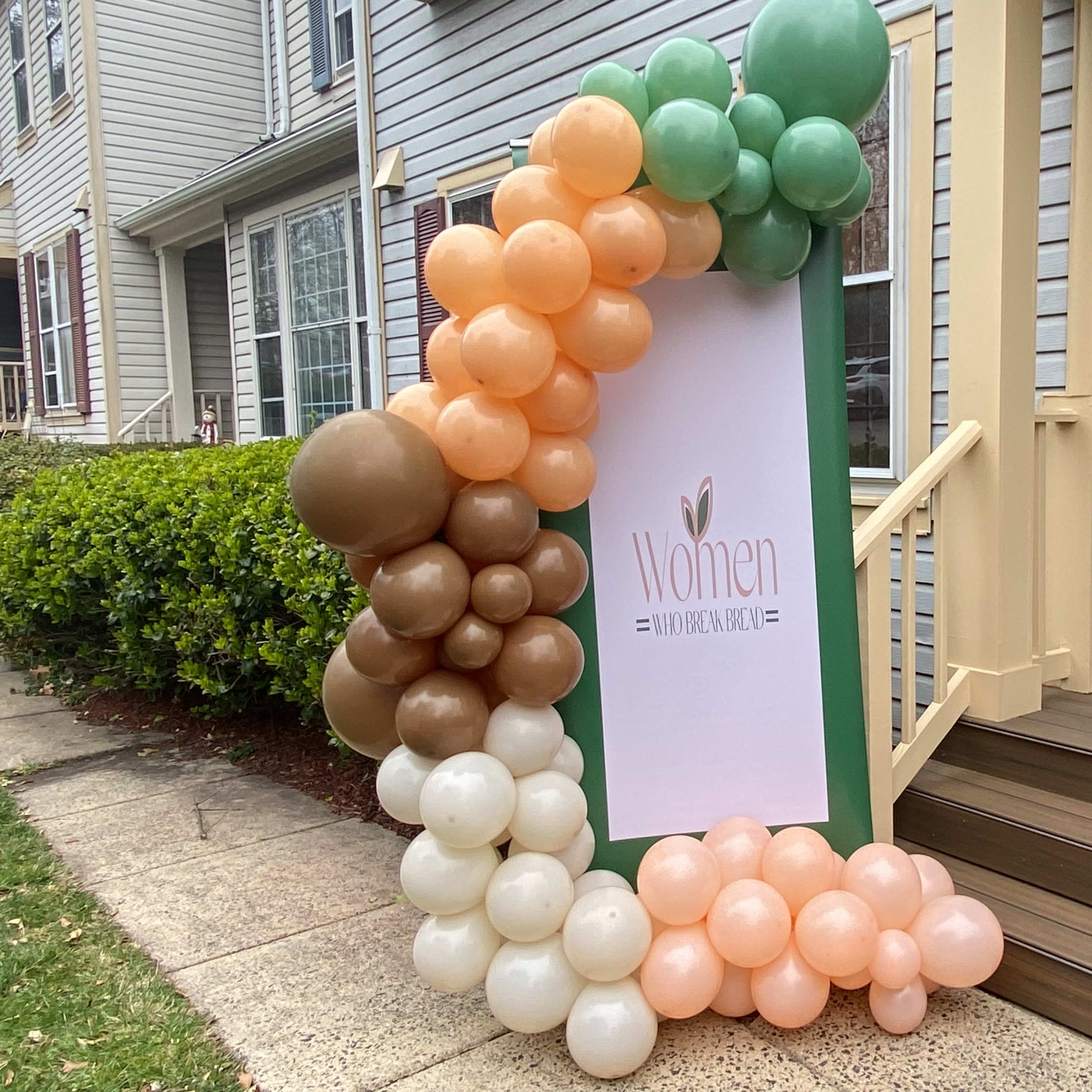 A decorative balloon arch in shades of green, peach, and brown frames a sign that reads "Women" at the entrance of a building.