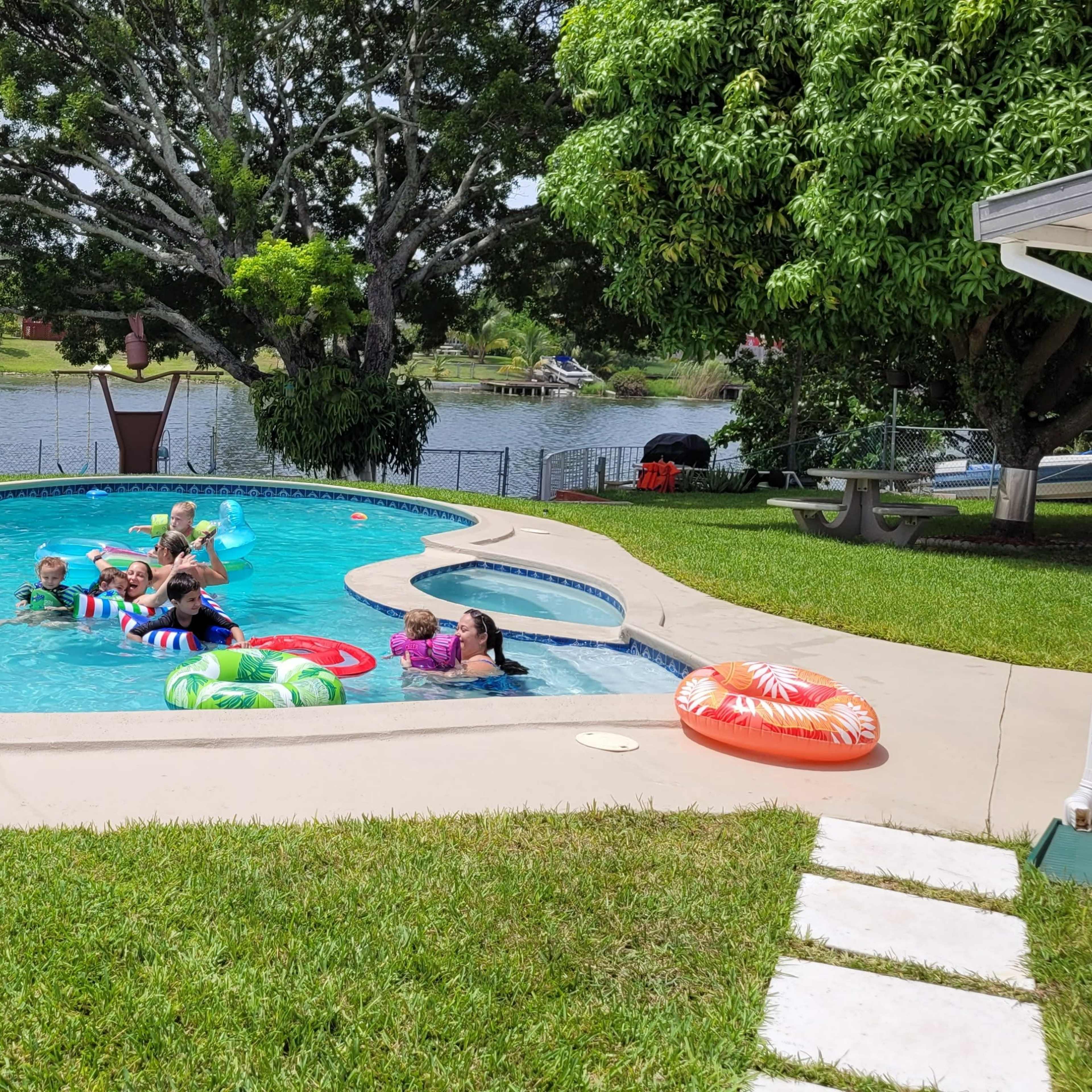 A group of people is enjoying a swimming pool with colorful floats next to a large tree and a lake in the background.