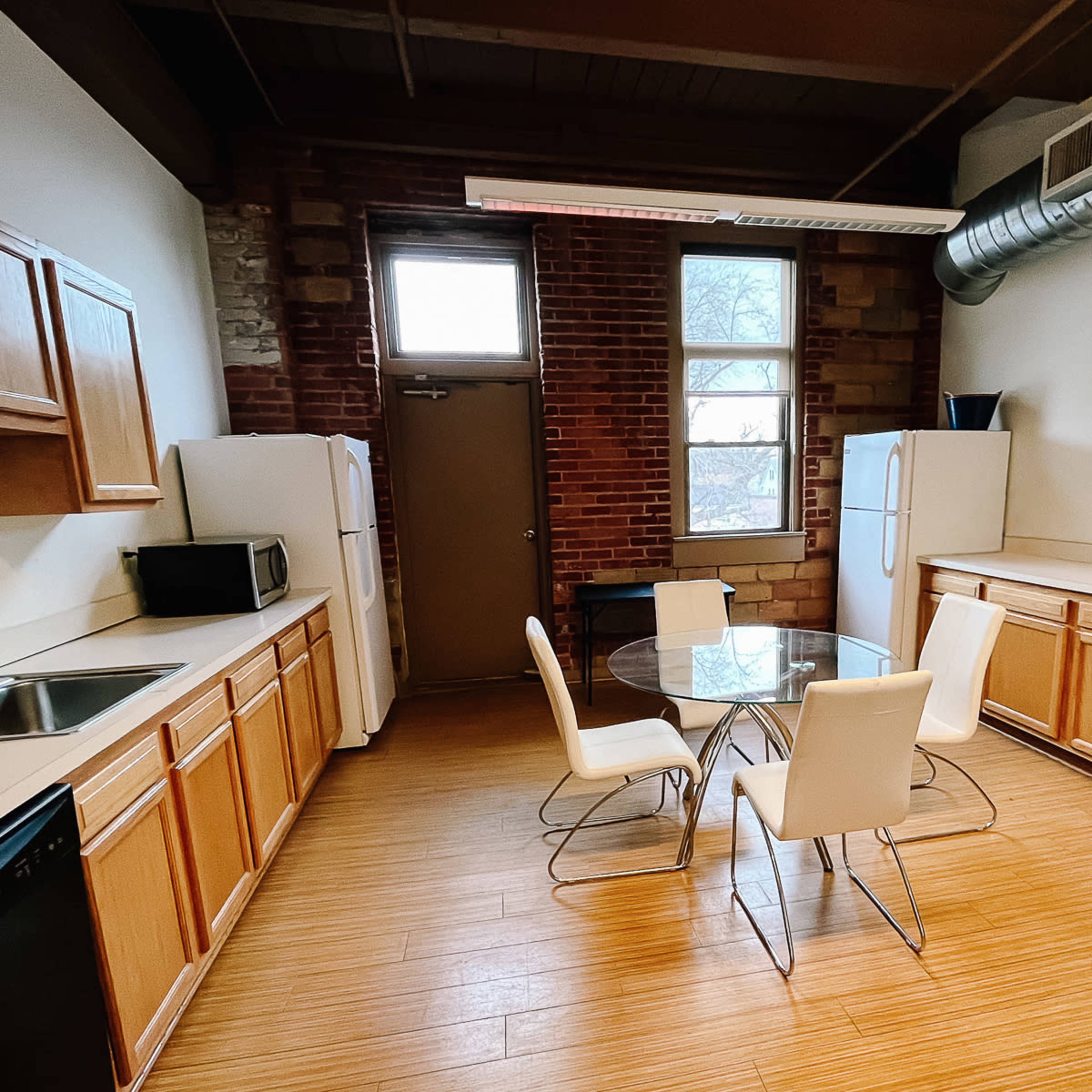 A kitchen with wooden cabinets, a round glass dining table surrounded by four white chairs, and two refrigerators against a brick wall.