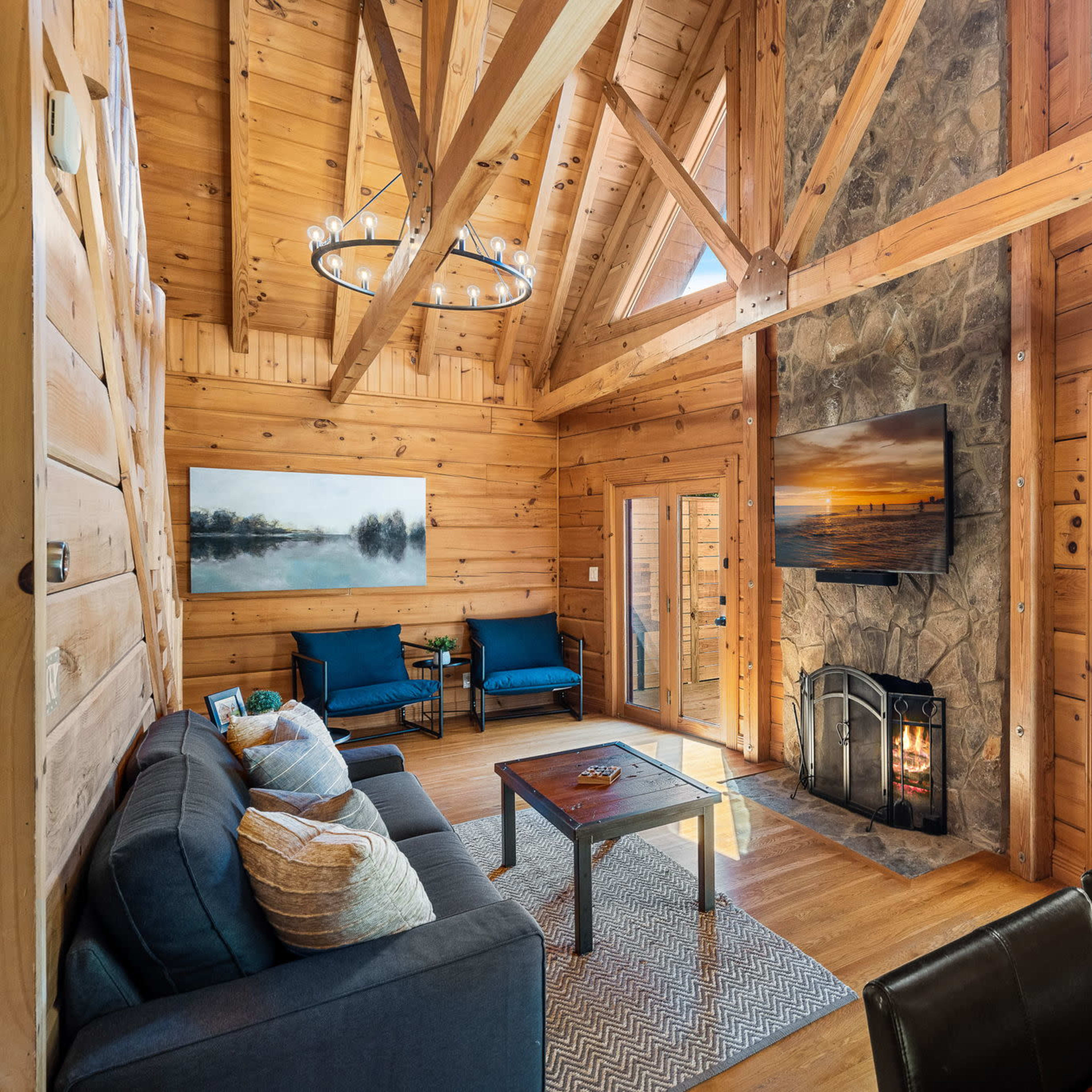 A spacious wooden cabin living room features a stone fireplace, a large window overlooking a deck, and a seating area with a sofa and chairs.