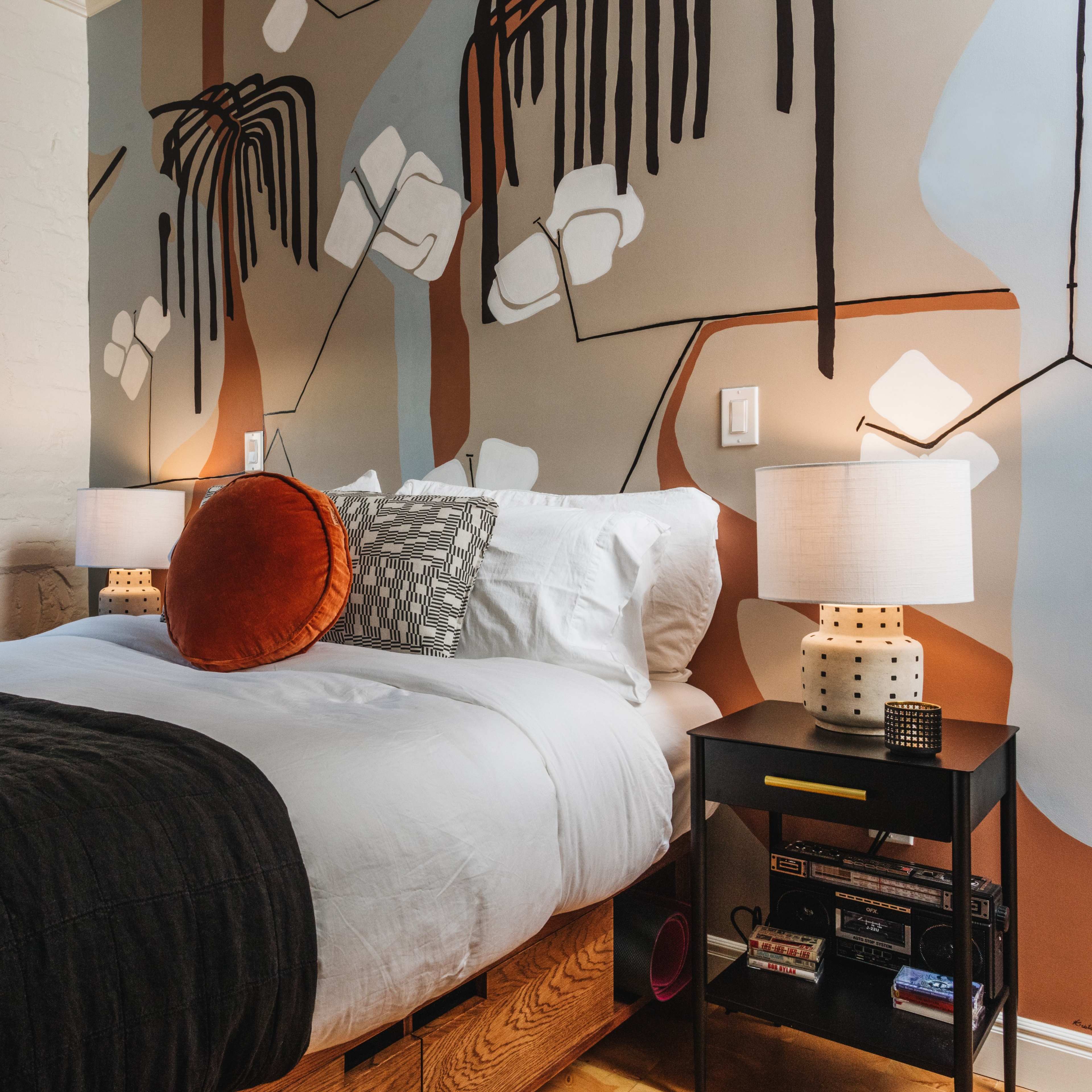 A neatly made bed with decorative pillows and a nightstand beside it, set against a stylized mural on the wall and a window with a view of an outdoor area.
