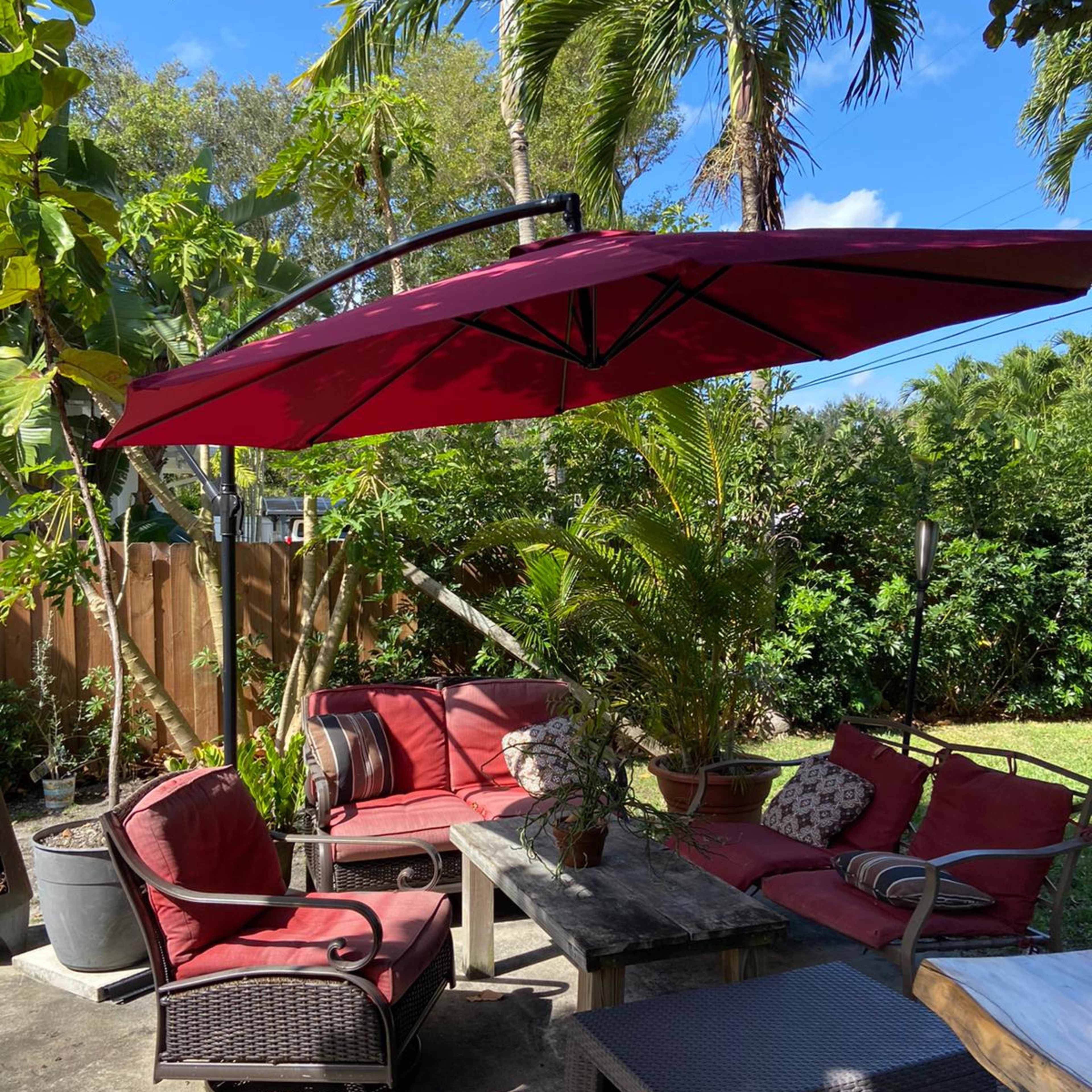A shaded outdoor seating area features red cushions on furniture surrounded by tropical plants and trees.