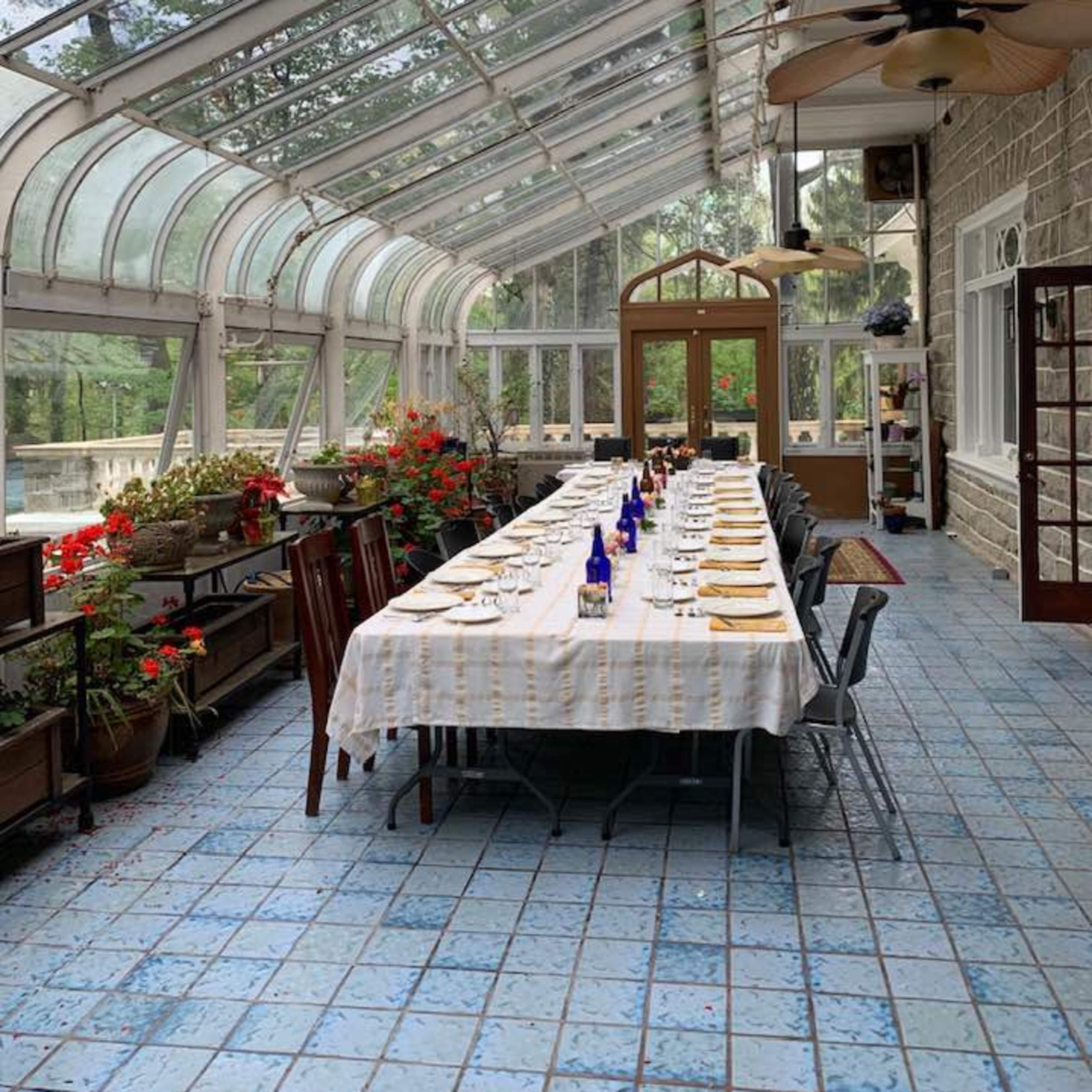 A long dining table is set under a glass-roofed structure with potted plants and flowers along the sides.