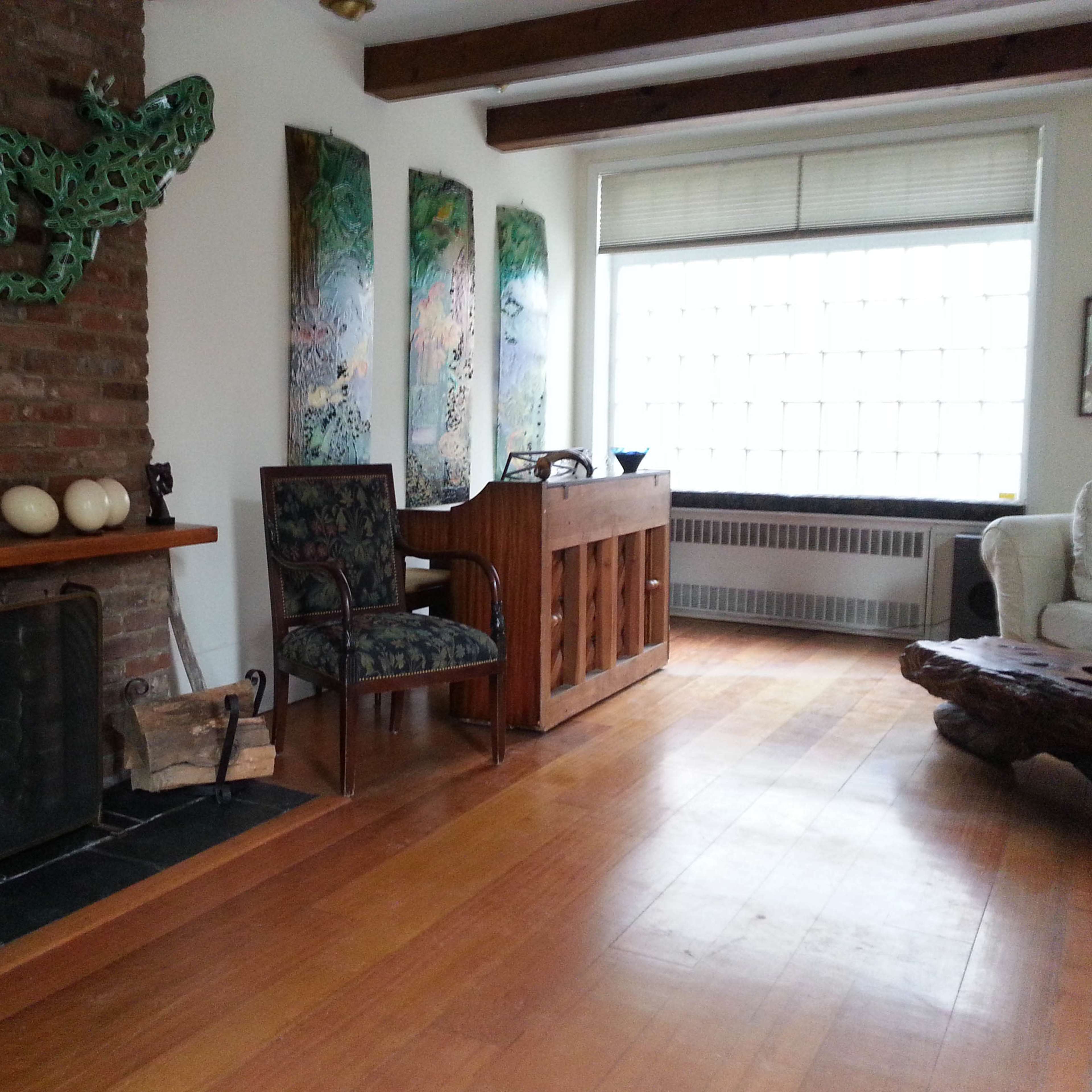 A living room with a brick fireplace, a green frog sculpture on the wall, wooden flooring, and paintings next to a window.